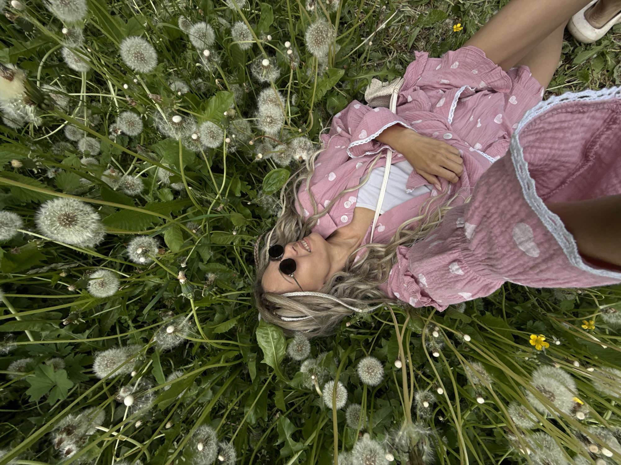 An overhead photograph captures a woman lying relaxed amidst a vibrant field of green grass and numerous white dandelion seed heads. She is positioned diagonally across the frame, dressed in a loose-fitting pink garment adorned with small white heart patterns and white lace trim, worn over a white top. Her long, wavy blonde and light brown hair is spread out, with some strands secured by a white band. She wears dark, round sunglasses and displays a joyful, wide smile, indicating a state of happiness and contentment. Her hands rest gently on her midsection. Scattered among the dandelions are small yellow wildflowers. A light-colored shoe is partially visible in the upper right corner, and a woven strap from a bag or purse can be seen near her right shoulder. The scene depicts a peaceful outdoor moment during the daytime. No text is visible in the image. The location is Rusanovo, Russia.