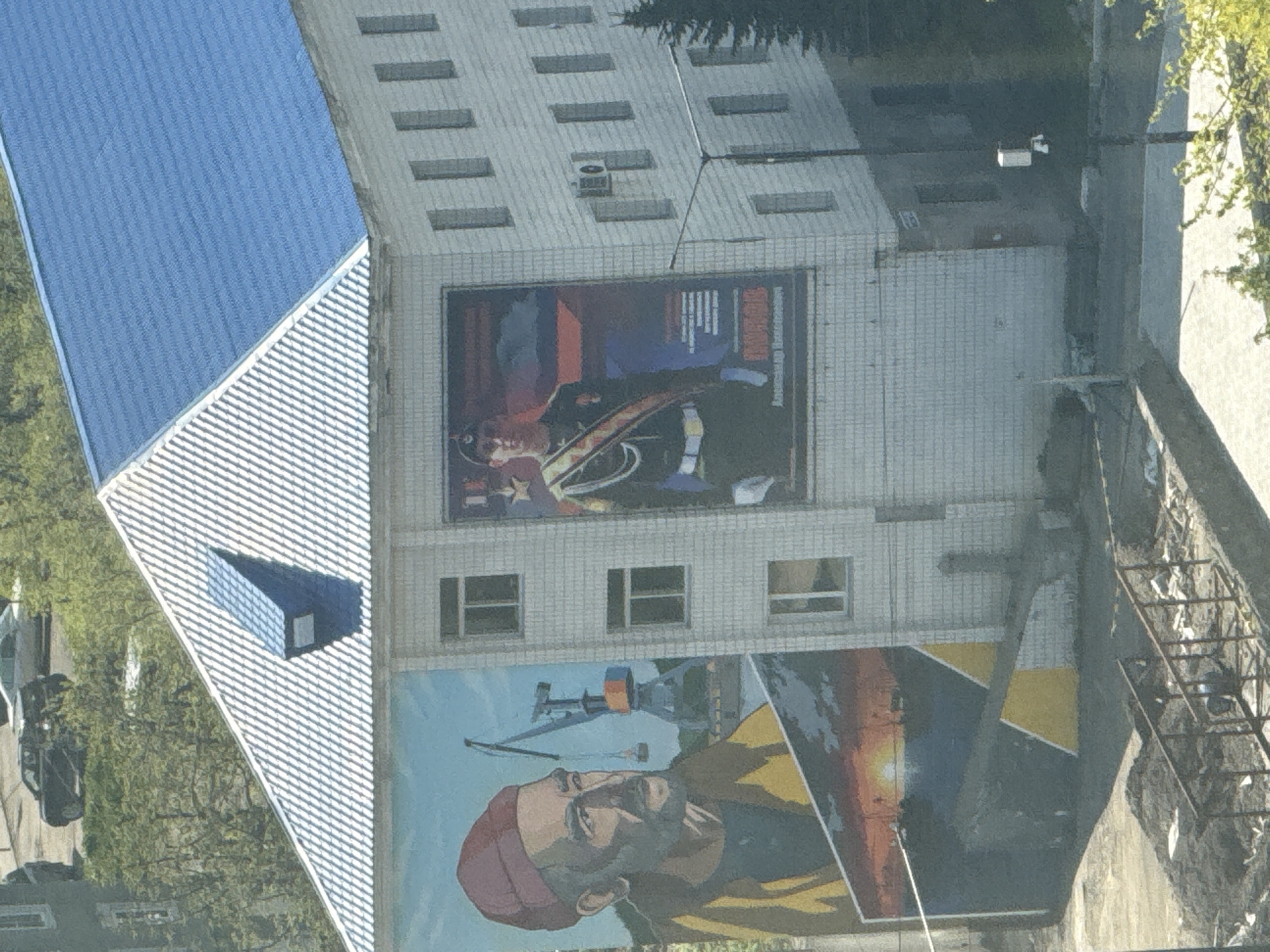 An image captured in Petropavlovsk-Kamchatsky, Russia, depicts a multi-story building featuring two large exterior murals and a distinct roof structure. The building's facade is constructed of light-colored square tiles, and it has multiple rectangular windows. The roof is bifurcated into two sections: the left section is white with a corrugated or diagonally patterned surface and includes a small blue dormer-like structure, while the right section is uniformly blue, also appearing corrugated.
On the left side of the building, a large mural portrays an older man with a beard, wearing a red beanie and a yellow jacket. He gazes towards the right. Behind him, an industrial port scene is depicted, featuring a large crane over a body of water. Below this, a segment of the mural illustrates a sunset over water with a silhouetted horizon. The lower part of the mural is characterized by geometric yellow and dark grey shapes.
On the right side of the building, a prominent vertical banner-style mural displays a young man in a dark green military uniform, complete with a white belt, a gold and red sash, and a dark cap bearing an insignia. Medals or ribbons are visible on his chest. The background is a blend of red, orange, and blue. Below the portrait, the name "ПАВЛОВ" (Pavlov) and "Александр Николаевич" (Alexander Nikolaevich) are clearly visible in Cyrillic script. An air conditioning unit is affixed to the building wall adjacent to this banner.
The foreground shows disturbed earth or gravel, possibly a construction area, with a low, rusty metal railing. Beyond this, a paved surface leads to a street light pole with an attached white security camera or sensor. In the background, beyond a line of green trees and bushes, a street is visible with several parked vehicles, including a black SUV and a silver sedan. Another building with a grey facade is partially visible on the far left. The sky overhead is clear and blue. The scene is illuminated by natural daylight.