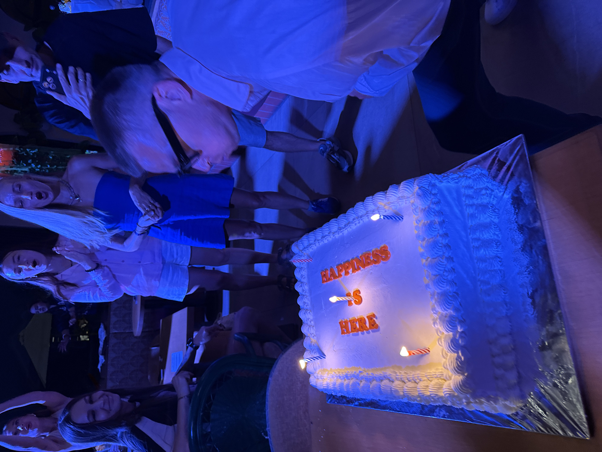 Here's a description of the image:
A high-angle shot showing a group of people around a birthday cake. The cake is square, white frosted, and has the words "HAPPINESS IS HERE" written in orange lettering. There are several candles lit on the cake. The people surrounding the cake appear to be celebrating and taking photos. The location of this event seems to be North Kuta, Indonesia. The atmosphere is festive, with a casual dress code among the attendees. The lighting appears to be a mix of natural and artificial. The specific environment, judging from the surrounding elements, looks like an outdoor event.