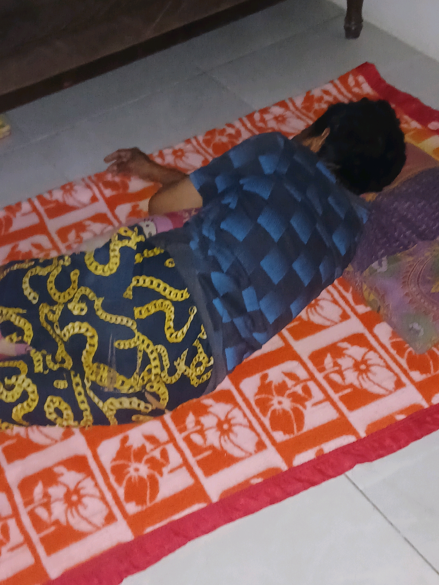 An overhead, medium shot captures a person lying on their stomach on a patterned mat on a tiled floor, near a wooden piece of furniture. The person is wearing a blue and black checkered t-shirt and dark blue pants with a yellow chain pattern. A pillow with a floral pattern is under their head. The mat has a red border and a white and orange floral pattern. The person is facing away from the camera, with their head turned to the left. The lighting is dim. It is unclear if this scene is in Gorontalo, Indonesia.