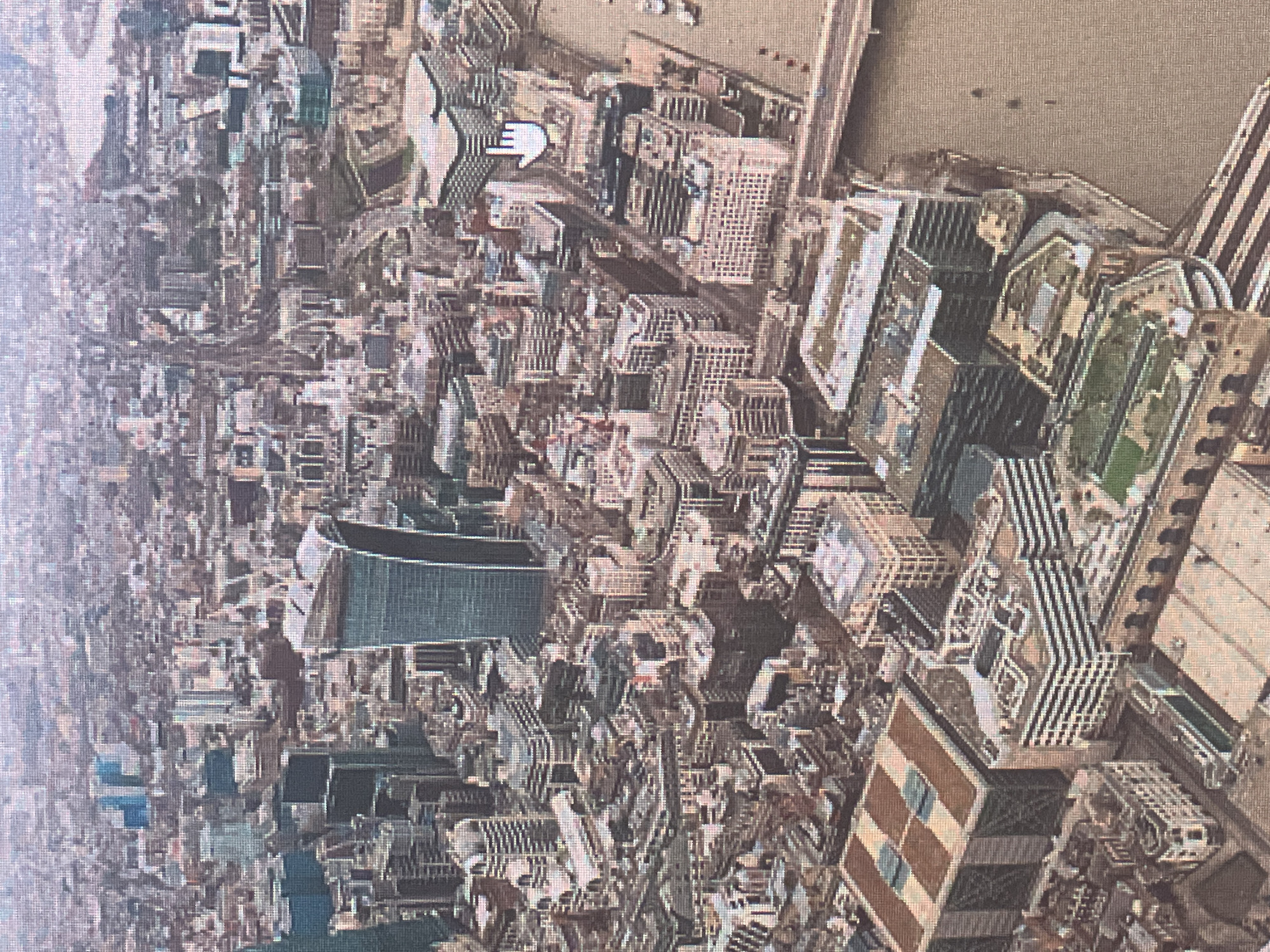 This is an aerial, slightly oblique photograph of a dense urban cityscape, dominated by numerous modern and older buildings. The most prominent structure is a distinctively curved, tall skyscraper with a dark glass facade, resembling a shard of glass. Surrounding this are many other buildings of varying heights and architectural styles, including office buildings with grid-like windows and some older, more traditional structures. In the foreground and to the right, a wide, muddy river runs through the city, with a bridge spanning it in the distance. Several boats are visible on the river. The overall impression is one of a bustling metropolis with a mix of contemporary and historical architecture, set against a hazy, overcast sky. There is no visible indication of any people or specific events occurring in the scene.