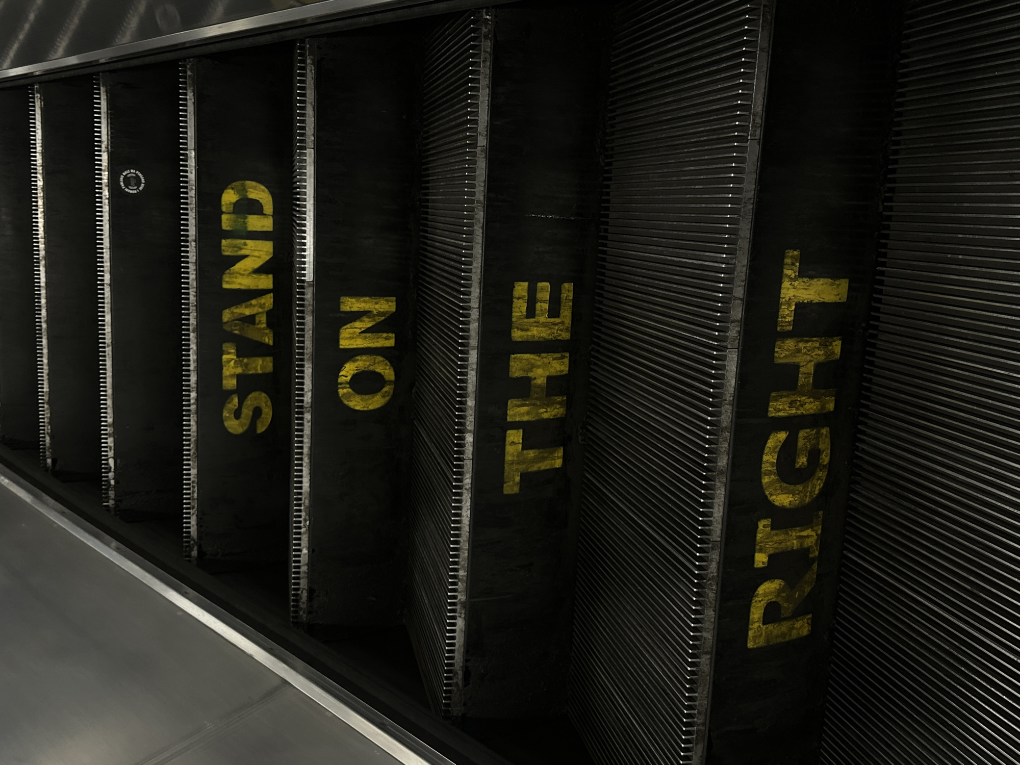 A low-angle shot depicts an escalator in the London Underground in the United Kingdom. The escalator steps are black, with yellow capital letters printed on them spelling out "STAND ON THE RIGHT." A small, round sign is present on the first step. The metal grates between the steps are visible. The escalator is made of metal and has a metallic silver trim.