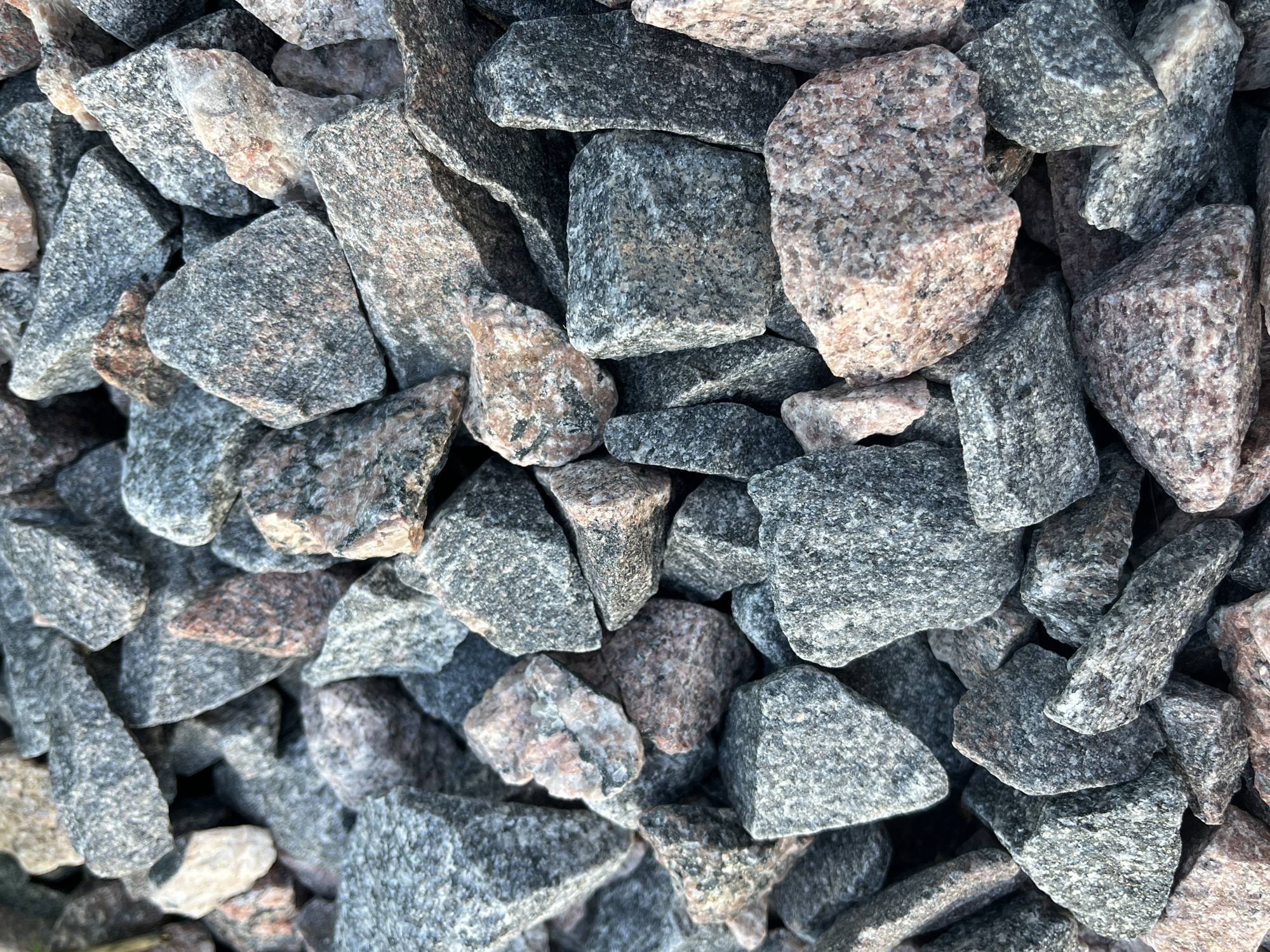 A close-up, overhead view shows a pile of rough, angular granite stones. The stones vary in size and color, with dominant hues of gray, black, and pink. The texture of the granite is visibly coarse, with individual mineral grains apparent. The stones are densely packed together, creating a textured surface with shadows cast in the crevices between them. There are no people, animals, or man-made objects present in the image. The scene is an outdoor, natural environment, likely a construction site or a landscape feature in Il'ino, Russia, given the context.