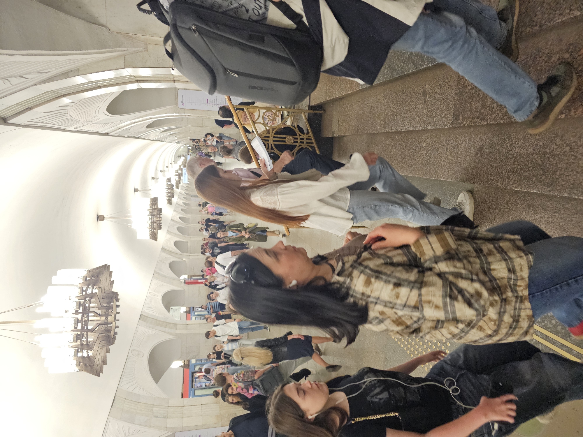 The image depicts a bustling scene within a grand, ornately decorated underground subway station, viewed from a slightly elevated perspective looking down a long, spacious hall. The station boasts high, vaulted ceilings adorned with intricate relief work and a series of ornate arches lining the walls. Large, multi-tiered chandeliers with geometric designs hang from the ceiling, casting bright, artificial light across the expansive space.
Numerous people are visible, engaged in typical commuter activities. In the immediate foreground, a person with long dark hair, wearing a plaid shirt and blue jeans, stands facing right. Below them, another person is partially visible from the chest down, wearing a dark top, blue jeans, and white earbuds. Further up, a person with long reddish-brown hair and a white top ascends a flight of stone stairs. Above them, a person carrying a black backpack also moves up the stairs.
Beyond the staircase, the platform extends into the distance, populated with more commuters. Some individuals are seated on benches lining the walls, while others stand or walk. A golden, decorative metal barrier or railing is visible near the top of the stairs. The stairs themselves are made of a speckled, textured stone material.
The architectural style, characterized by its grand scale, elaborate chandeliers, and detailed relief work on the ceilings and arches, is highly characteristic of historic stations within the Moscow Metro system. A white sign with some unreadable purple text is partially visible on a wall. The scene is brightly lit by artificial light, making the specific time of day or weather outside indeterminate.