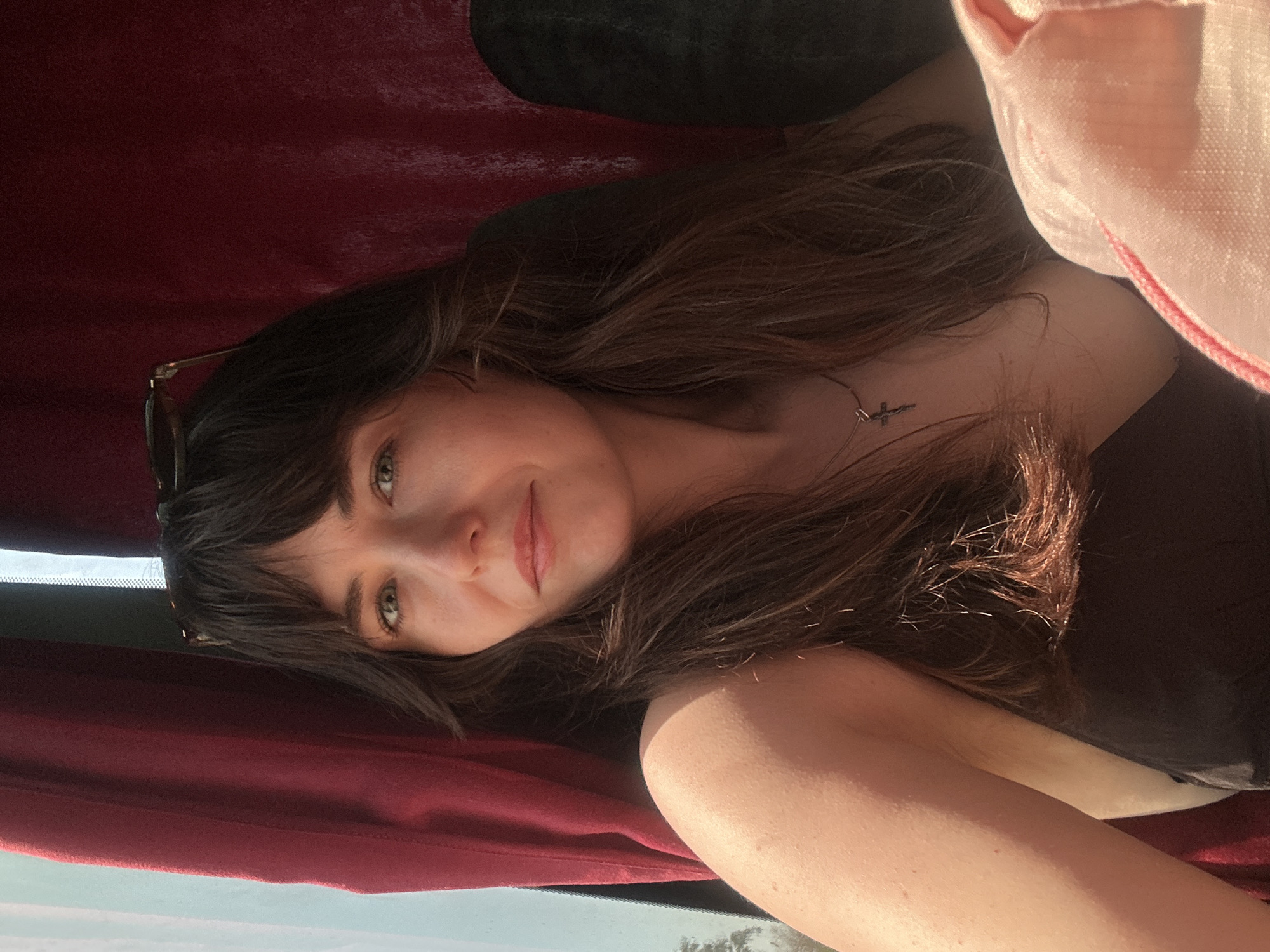 A medium shot depicts a young woman with long, brown hair and light eyes, positioned at an angle. She is wearing a dark-colored top and a delicate necklace with a small pendant. Sunglasses are placed on top of her head. The background includes a dark red curtain or fabric and possibly a headrest. A pink bag or fabric is visible on the right side of the frame. The lower portion of the image shows what appears to be a window with a view of a bright, outdoor scene.
The location is potentially Alanya, Turkey based on the prompt, although there is nothing visible in the image to confirm this. No actions or interactions are depicted. The environment suggests an indoor space, possibly a vehicle or a room with specific decor.