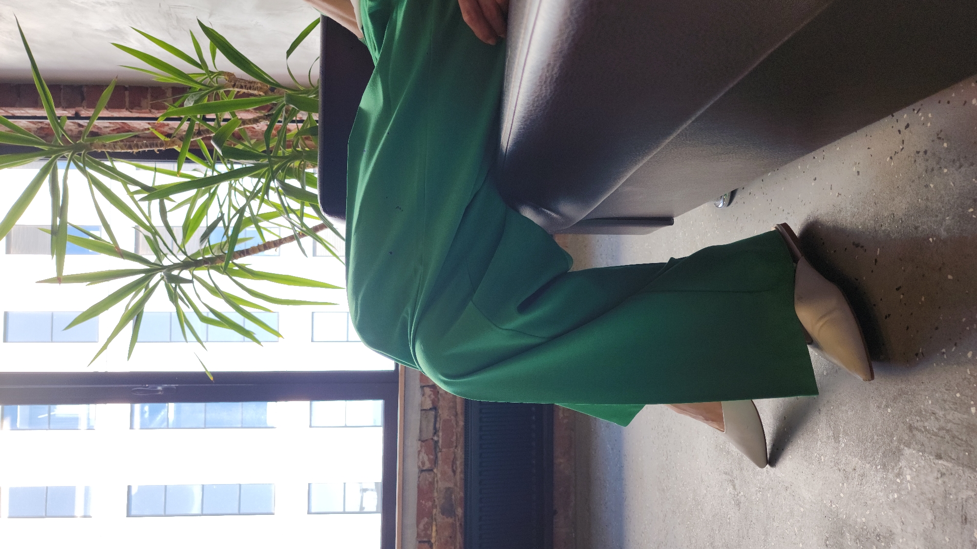 The scene presents an individual seated on a grey upholstered bench or sofa. They are wearing vibrant green wide-leg trousers paired with beige or light-colored pointed-toe heeled mules. A potted yucca plant, with its characteristic spiky green leaves, is positioned near the seating area.
A window is visible in the background, revealing an exterior with a grid-like pattern of rectangular panes, suggesting a modern or renovated building. A section of brickwork is also noticeable, possibly part of an interior or exterior wall.
The location is Moscow, Russia, as inferred from the architectural style observed outside the window. The interior design suggests an urban environment.