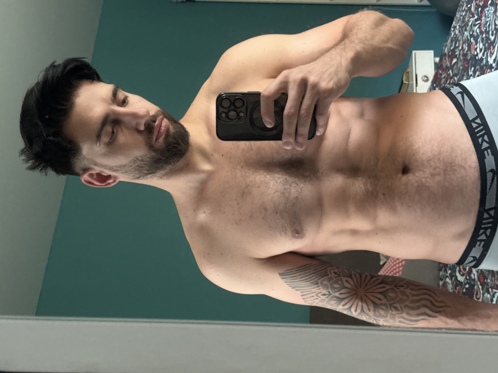 The image is a mirror selfie of a man, taken in an indoor setting, possibly in Sofia, Bulgaria. The original image is rotated 90 degrees counter-clockwise. The man has dark, medium-length hair with faded sides and a full dark beard. He is bare-chested, revealing a lean and muscular physique. He is looking downwards, towards his reflection or the phone in his hand, with a serious or thoughtful expression.
He holds a black smartphone with a prominent multi-lens camera system in his right hand, covering part of his chest. He is wearing light grey boxer briefs with a black waistband that clearly shows the brand name "NIKE" in white letters. His left forearm is adorned with a detailed black tattoo, featuring a central mandala or floral design surrounded by radial and wave-like patterns.
The background consists of a solid teal-colored wall on the upper left side. On the right, behind the man's lower body, a multicolored patterned fabric, possibly bedding or a curtain, with shades of red, black, and white, is visible. A small white electrical outlet or wall fixture can also be seen against the patterned fabric. The lighting suggests an indoor environment.