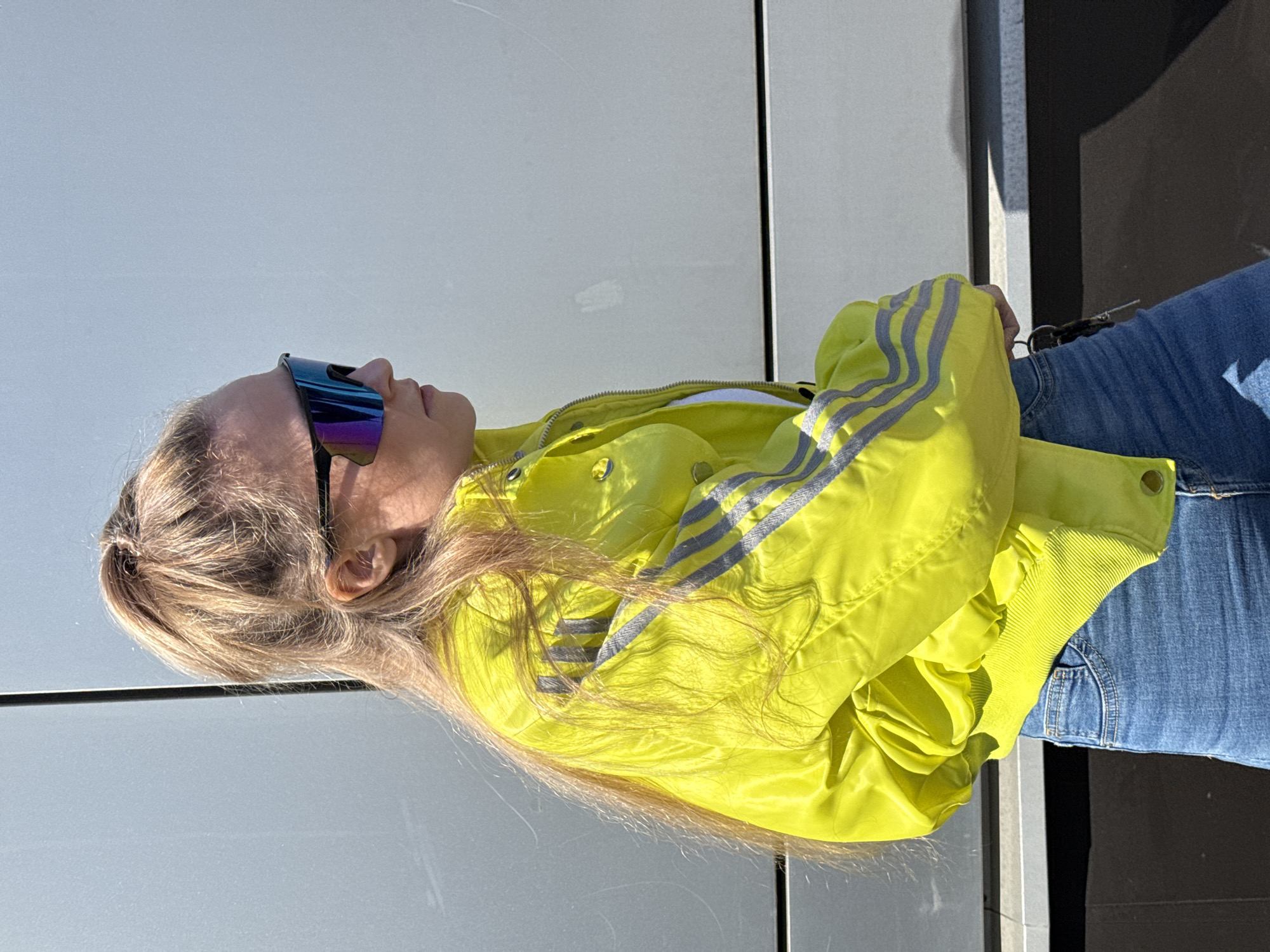 A female individual stands in profile, facing right, outdoors in Izhevsk, Russia. She wears mirrored, multi-colored wrap-around sunglasses with black frames. Her light-colored hair is styled in a high ponytail. She is dressed in a neon yellow bomber jacket, featuring three parallel grey stripes on the sleeves and ribbed cuffs/hem, worn over a white undergarment. Blue denim jeans cover her lower body. A set of keys is attached to her left hip area. The background is a large, light grey, reflective surface, possibly a vehicle or building wall, exhibiting minor scratches. Direct sunlight illuminates the scene, casting shadows, with a darker horizontal surface visible at the lower frame edge.
