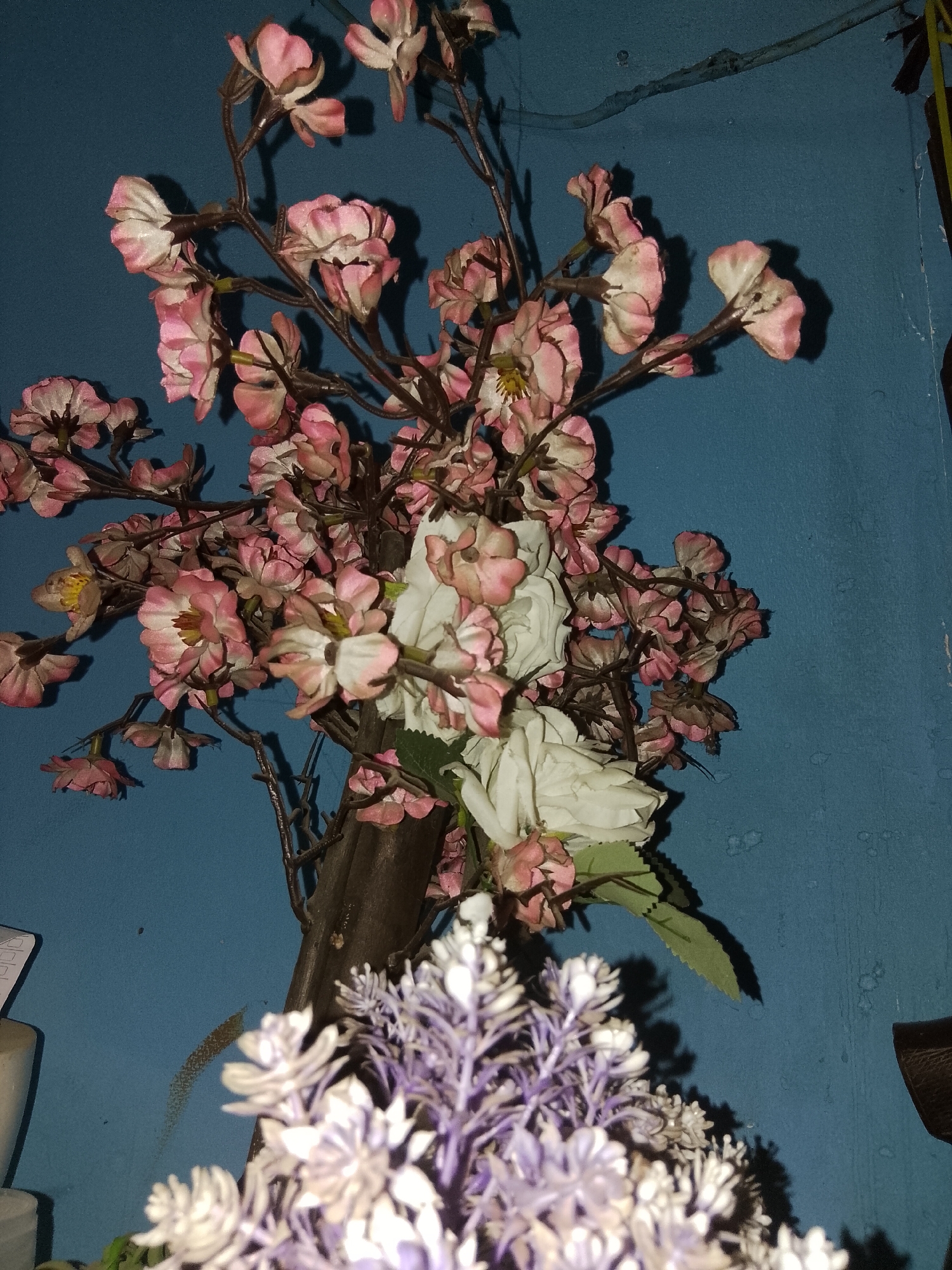 The image displays a close-up view of artificial flowers against a textured blue wall. The primary focus is a cluster of pink and white artificial flowers, appearing to be cherry blossoms or similar delicate blooms, attached to dark, twig-like stems. Some of these flowers show detailed texture and color variations, with hints of yellow in the centers. Interspersed among the pink and white flowers are larger, white artificial roses. In the foreground, out of focus, is a cluster of smaller, light purple and white artificial flowers, resembling lavender or baby's breath. The blue wall behind the flowers has a slightly mottled appearance, with subtle variations in color and some circular marks. The lighting in the image is dim, casting soft shadows. There are no people or animals visible in the image, and no overt actions are taking place. The scene suggests an indoor decorative arrangement. The mention of Bencongan, Indonesia, suggests this photograph was taken in that specific location, likely within a home or a similarly private space, given the nature of the subject matter.