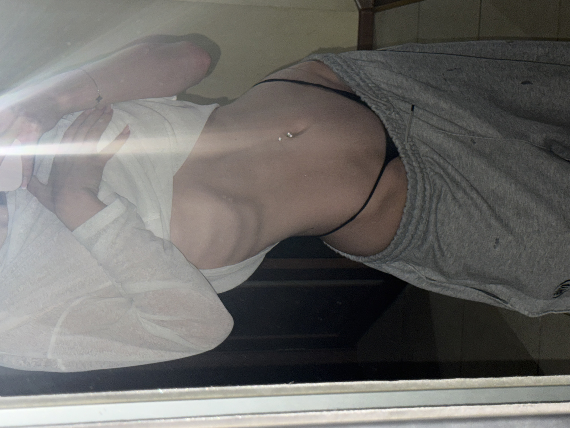A person is captured in a mirror selfie, posing in what appears to be a dimly lit room. They are wearing a sheer, long-sleeved white top, pulled up to expose their midriff, and grey sweatpants. A black thong is visible at the waistline of the sweatpants. The person has a navel piercing with two small, clear gems. Their left arm is resting on their stomach, and their right hand is touching their chest. A thin silver bracelet with a clover charm is worn on their right wrist. The background suggests an indoor setting with a door and a portion of furniture visible. The image does not contain any specific landmarks or features that would allow for localization to Khimki, Russia.
