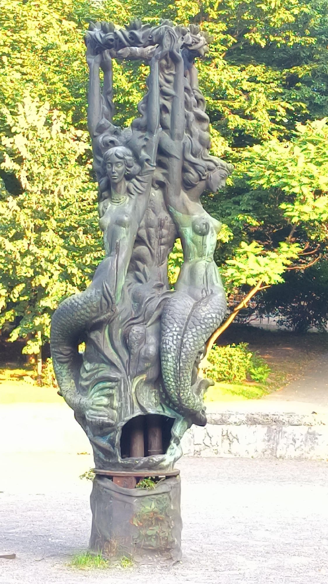 The image shows a bronze sculpture of mermaids with intertwined tails and human upper bodies, holding a floral wreath above their heads. The sculpture is set against a backdrop of lush green trees. The base of the sculpture is a dark, rough material, with some greenery growing around it. The ground in the foreground is a light gray gravel. The sculpture is located in Lviv, Ukraine, specifically in Ivan Franko Park, near the Ivan Franko National University.
