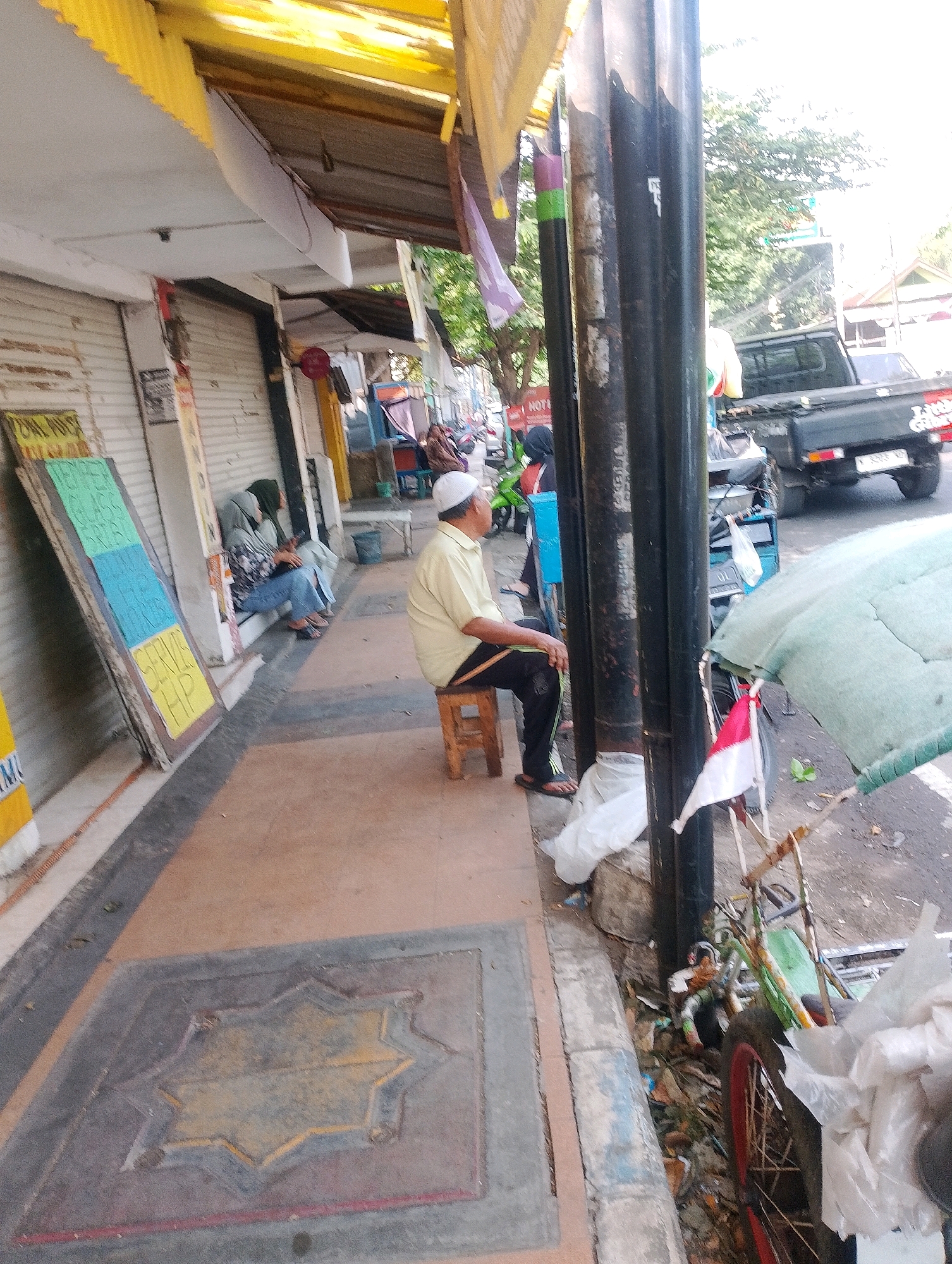 The image captures a street scene in Probolinggo, Indonesia, likely a commercial area with storefronts. In the foreground, a man wearing a white cap and a light yellow shirt sits on a wooden stool, facing away from the camera. He is dressed in black pants and sandals. To his left, on the sidewalk, are several closed storefronts with roll-up metal doors. A slanted board with colorful signage, possibly advertising services, leans against one of the storefronts. Further back, two individuals are seated on a step, one wearing a headscarf and jeans, and the other also partially obscured. The street is visible in the background, with a black pickup truck parked on the right side of the frame. Next to the pickup truck is a bicycle with a green frame and a red rim, and an umbrella or canopy made of green fabric. The scene suggests a bustling but somewhat cluttered environment, with debris visible on the ground near the bicycle. The overall impression is of a typical Indonesian urban street during daylight hours.