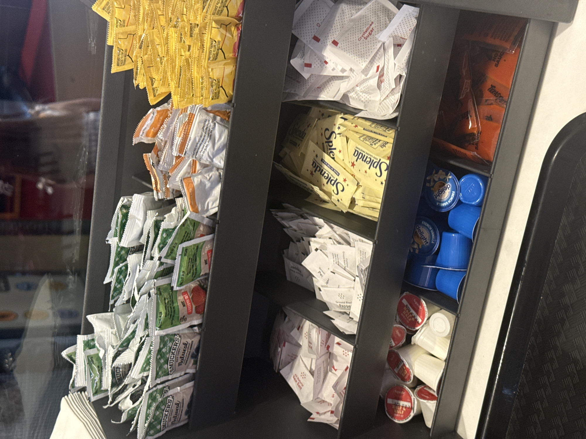 The image shows a condiment station, likely in a fast-food restaurant, possibly located in or near Mountain Park, Georgia, United States. The station is organized into several vertical sections. From left to right, the compartments contain: packets of yellow mustard, orange-labeled sauces, green packets of Smucker's Natural jam and other spreads. The middle section holds packets of white crystallized sugar, yellow Splenda artificial sweetener, and additional packets of white powder, possibly salt or pepper. The right-most section holds Tangy hot sauce, blue-lidded cups of creamer and half-and-half in individual containers. The station is made of dark gray material with a metallic accent. A black tray is visible on the right edge of the frame. The surroundings appear to be interior and somewhat dimly lit.