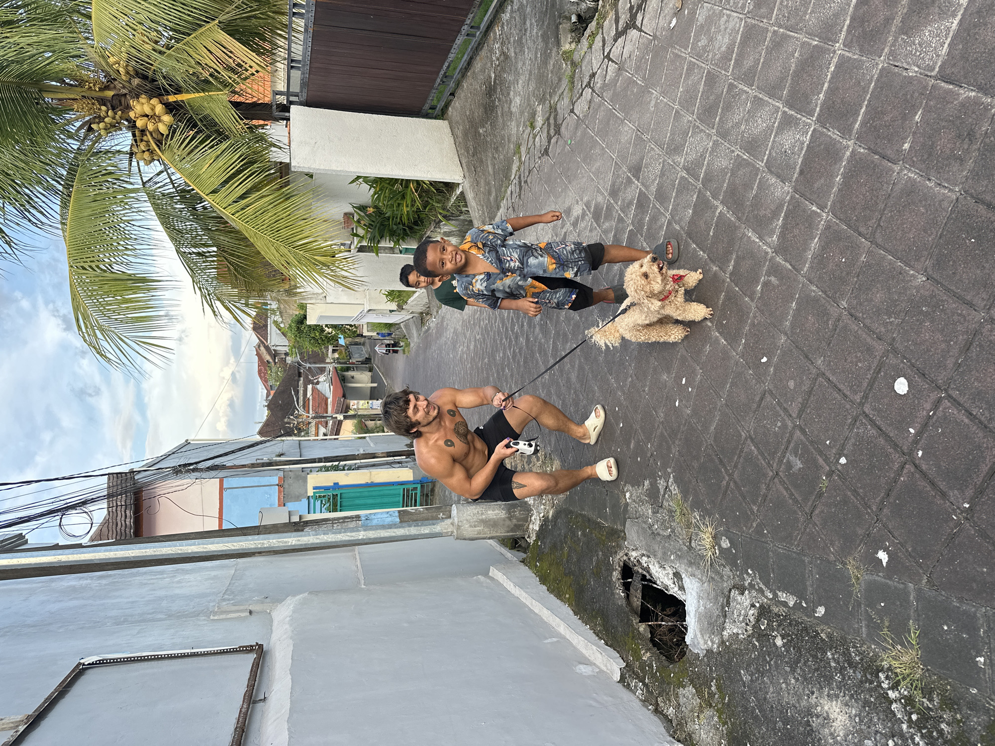 In Dalung, Indonesia, a shirtless man with dark hair, tattoos on his chest and left thigh, and wearing white sandals is crouched on a grey-bricked path, holding a dog leash. The leash is attached to a medium-sized beige poodle wearing a red collar, which is lying on the path. Two young boys, one wearing a patterned blue and grey shirt and black shorts, are standing to the right of the dog, closer to the end of the path. A third boy is partially visible behind them.
To the left, there is a light grey wall. To the right, there is a drainage hole in the path near the wall. A coconut tree is partially visible at the top-left of the image. Overhead, a jumble of power lines is visible. Buildings and rooftops can be seen in the background.