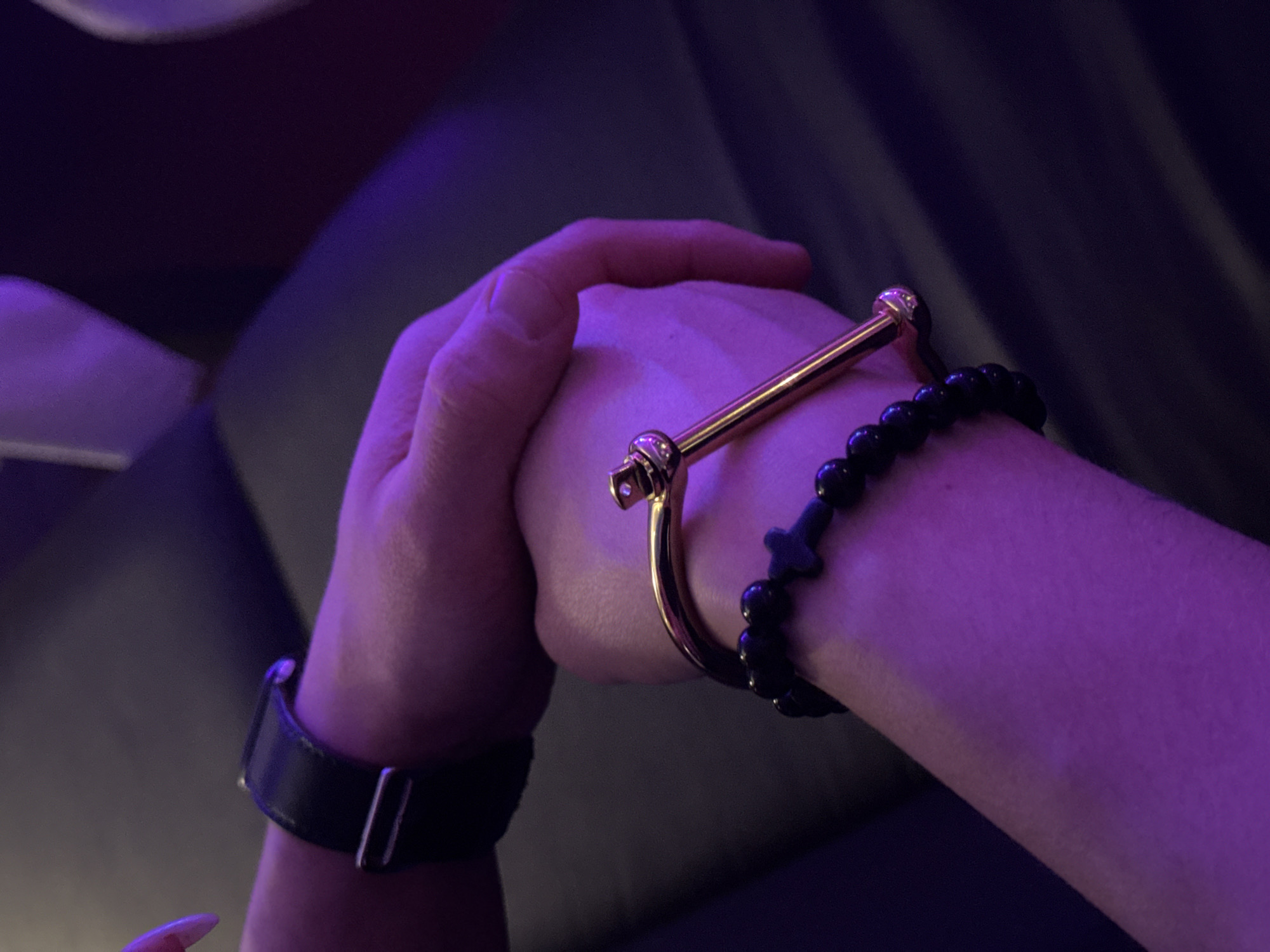 Two hands are depicted in a close-up, dimly lit scene under strong purple illumination. The upper hand, wearing a black watch strap with a silver-colored buckle on its wrist, gently holds the forearm of the lower hand. The lower hand's wrist is adorned with two bracelets: one gold-colored, tubular in design with a prominent screw-head-like closure featuring a small, clear stone, and another composed of black beads, including spherical beads and a single cross-shaped bead. The background is dark and out of focus, showing indistinct shapes suggestive of indoor furniture. The precise location within the city of Chaikovsky, Russia, is not discernible from the provided visual information.