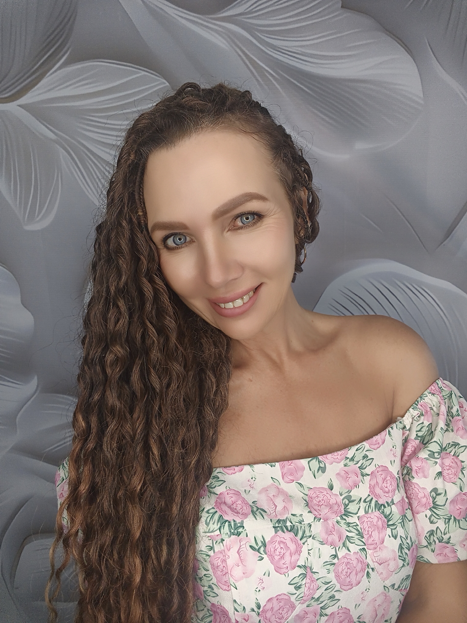 A medium shot photograph features a Caucasian woman with long, curly brown hair, blue eyes, and fair skin. She is smiling and looking towards the camera. The woman is wearing an off-the-shoulder dress with a floral pattern of pink roses and green leaves. The background is a gray, textured wallpaper with a floral motif. The lighting is bright, illuminating the woman's face and upper body.