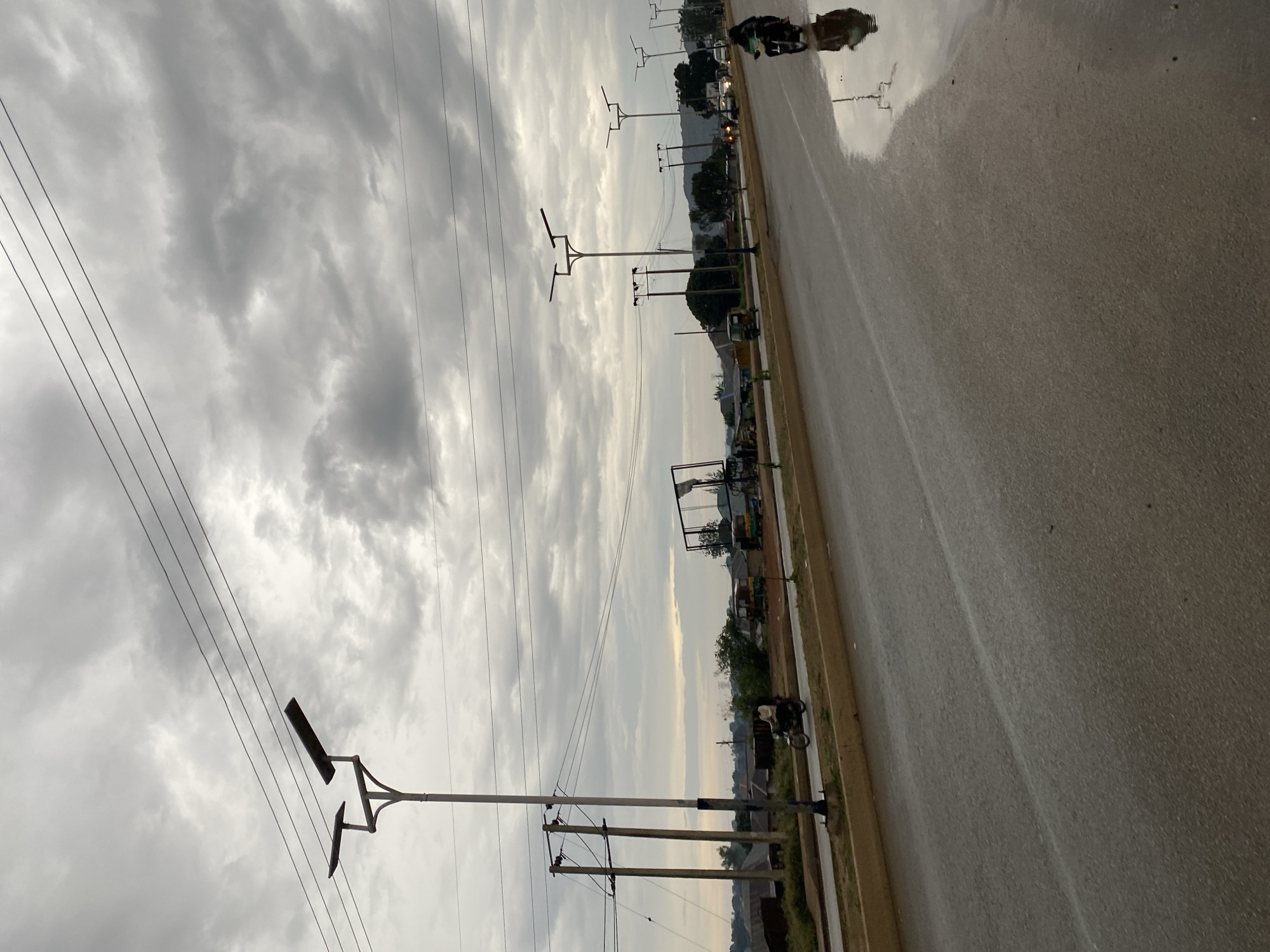 The original image, which is rotated 90 degrees clockwise, presents a subdued street scene in Bukuru, Nigeria. The scene is dominated by a heavily overcast sky filled with dense, grey clouds, suggesting recent rain or a very cloudy day. The wide paved road in the foreground is wet and highly reflective, mirroring the sky and distant elements, particularly on the right side.
Along the left side of the road, several utility poles support multiple power lines that stretch across the frame. Modern streetlights, some featuring double, angular light fixtures, are also prominent. On the right side, the wet road surface clearly reflects a person riding a motorcycle or scooter. Further into the distance along the right, other faint vehicle shapes with illuminated headlights are visible, alongside what appears to be a "Keke Napep" (auto-rickshaw). Buildings, structures, and some patches of greenery line the road in the background, characteristic of a semi-urban environment.
The overall lighting is diffused, indicating either early morning, late afternoon, or a dark, overcast day. The wet road is a key detail, implying recent rainfall. No visible text is discernible in the image. The general atmosphere conveyed is calm and quiet.