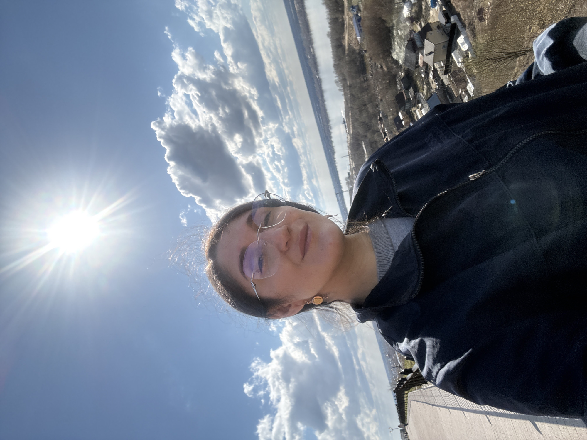 A selfie-style photo captures a young woman standing outdoors under a bright, sunny sky, offering a panoramic view of a landscape identified as Tobolsk, Russia. The image is oriented vertically with the person on the right side of the frame, facing slightly left towards the camera.
The main subject is a woman with dark hair, wearing glasses, a grey sweater, and a dark blue or black zipped jacket. She has a subtle, pleasant expression. A distinctive gold-colored circular earring is visible on her right ear.
The background is dominated by a vast blue sky with scattered, fluffy white cumulus clouds. A brilliant sunburst effect emanates from the upper left quadrant of the frame, indicating the sun's position. Below the horizon line, a wide, calm body of water, likely the Irtysh River, stretches across the middle distance. Beyond the river, a distant shoreline with a faint treeline is visible.
To the right of the river, in the mid-ground, lies a town or settlement with various houses and structures nestled among bare trees, suggesting a season like early spring or late autumn. The woman appears to be on an elevated pathway or viewing platform, with a dark railing visible along the right edge of the foreground. The weather is clear and sunny, indicative of midday or early afternoon, with a vast outdoor setting. There is no visible text in the image.