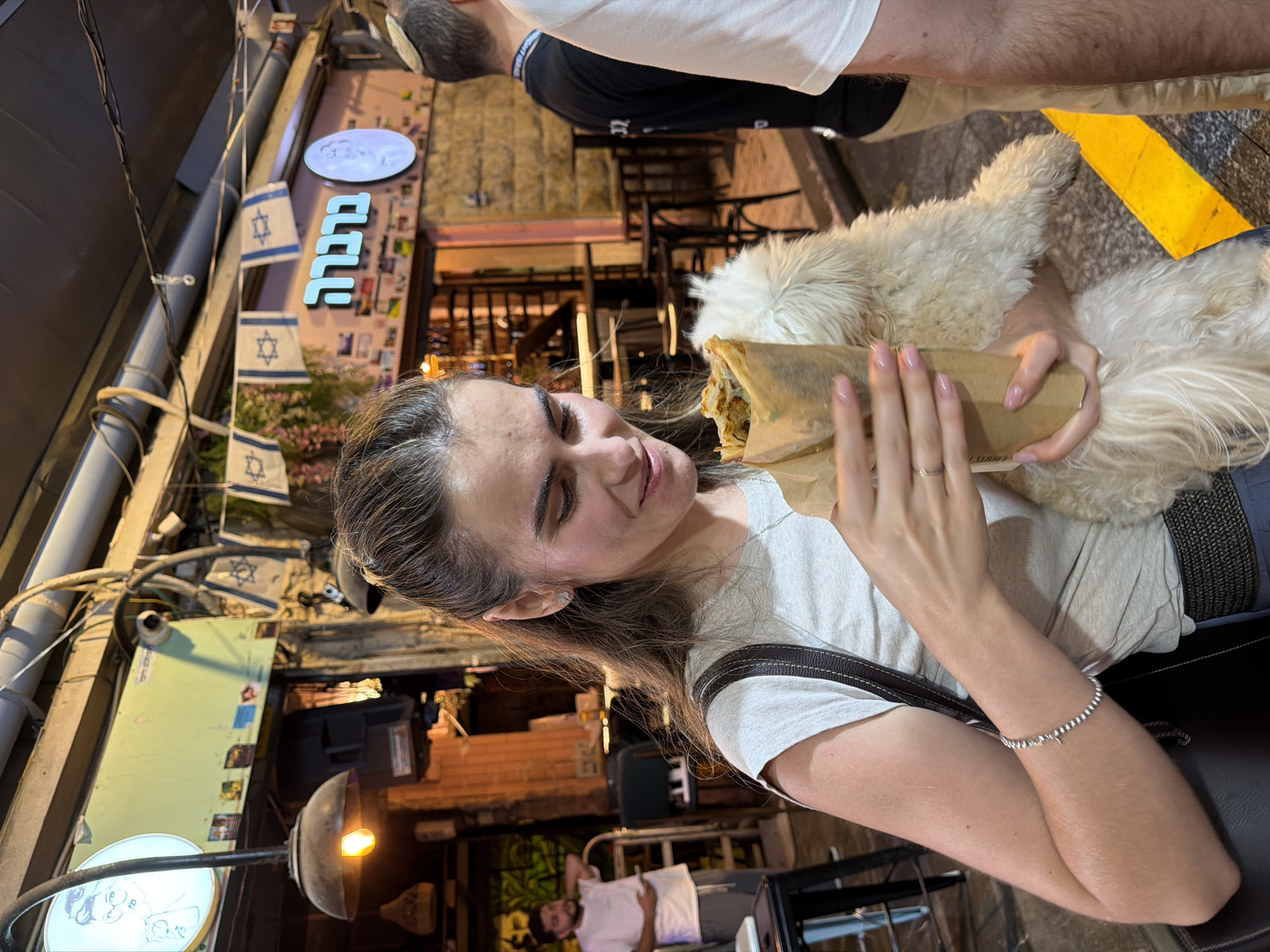 In a street scene at night, likely in Jerusalem, Israel, a young woman with her hair tied back is holding a dog and a food item wrapped in brown paper. She is wearing a light-colored t-shirt and has a silver bracelet on her left arm. The dog, a fluffy white breed, is nestled in her arms. Behind her, a storefront displays neon Hebrew signage that reads "ברבורה". Israeli flags are hung along the street. In the background, another man in a white t-shirt is visible, and further back, a man in a black shirt and another individual in a white t-shirt are present. The environment appears to be an outdoor dining or market area with tables and chairs set up. The lighting suggests a warm, evening atmosphere.