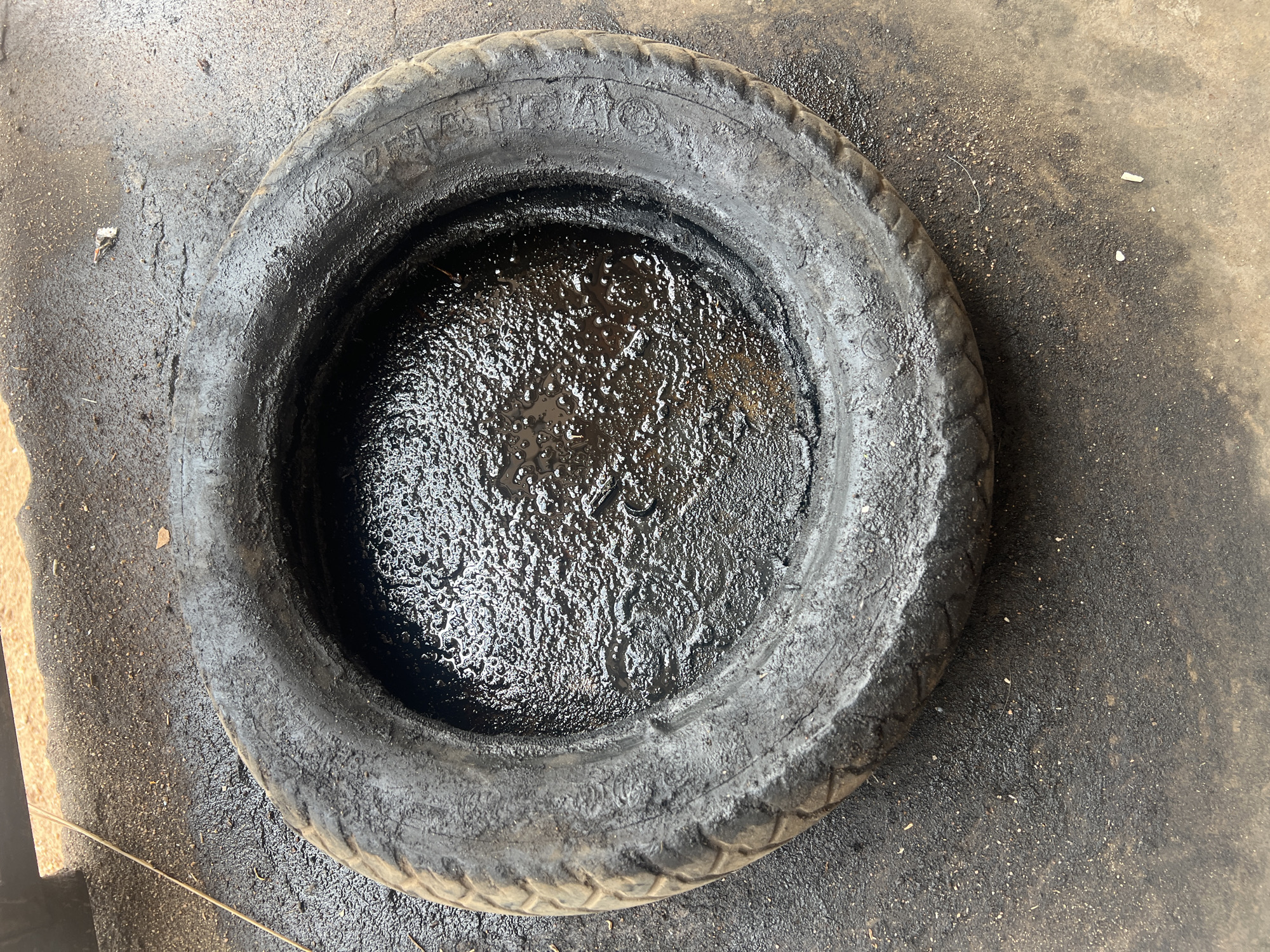 A used tire lies on a dirt and asphalt surface in Jalingo, Nigeria. The tire is filled with dark, viscous liquid, likely oil or waste. The ground around the tire is also stained with the same dark substance and scattered with small pebbles and debris. The overall impression is one of a neglected area, possibly a workshop or a disposal site. No people are visible in the image.