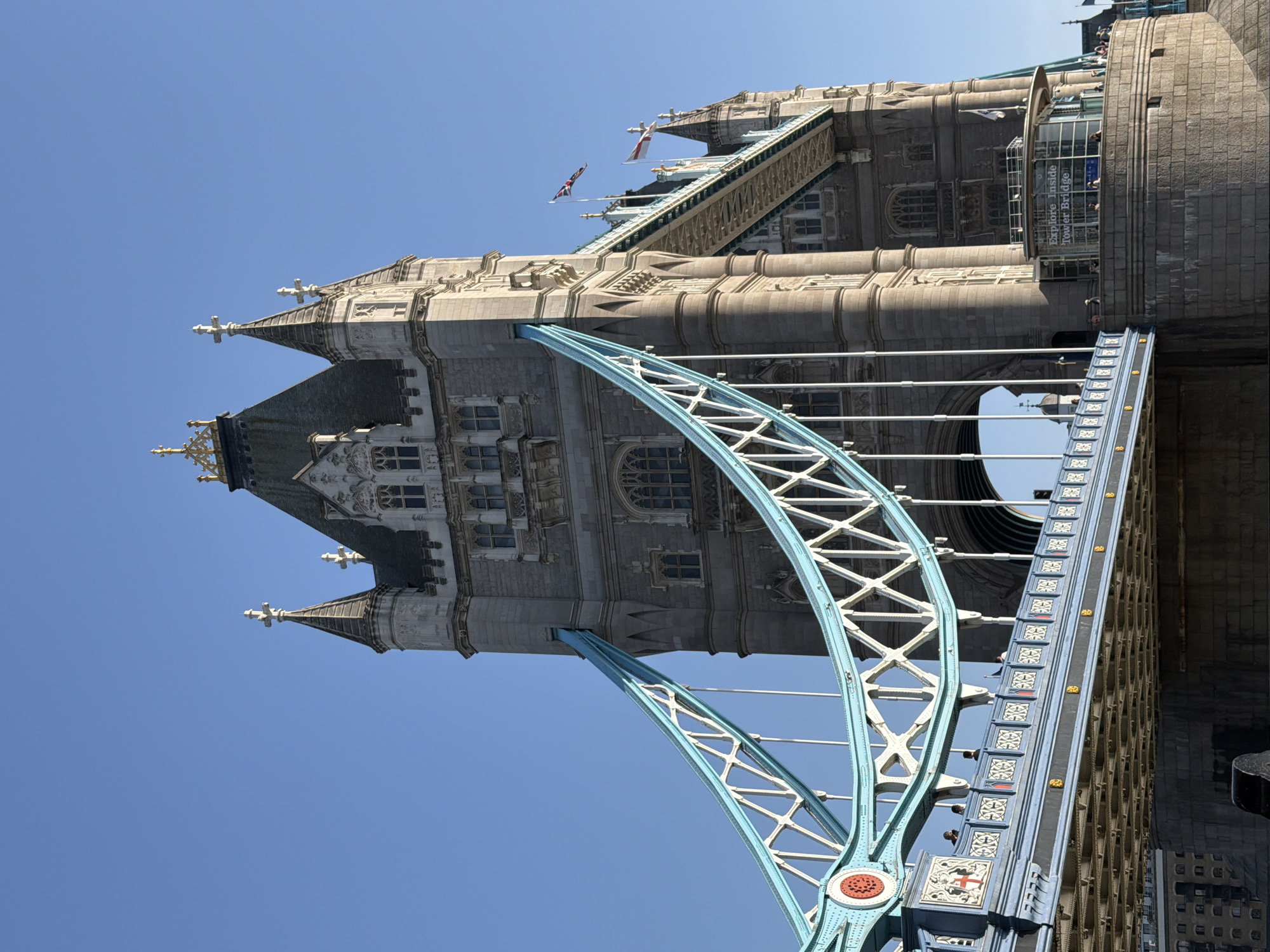 The image shows a portion of the Tower Bridge in London, United Kingdom, tilted at an angle. The architectural details of one of the bridge's towers are prominently visible against a clear blue sky. The bridge's distinctive steel framework, painted in light blue, contrasts with the gray stone of the tower. The image captures the structure from a low vantage point, emphasizing the bridge's height and design.