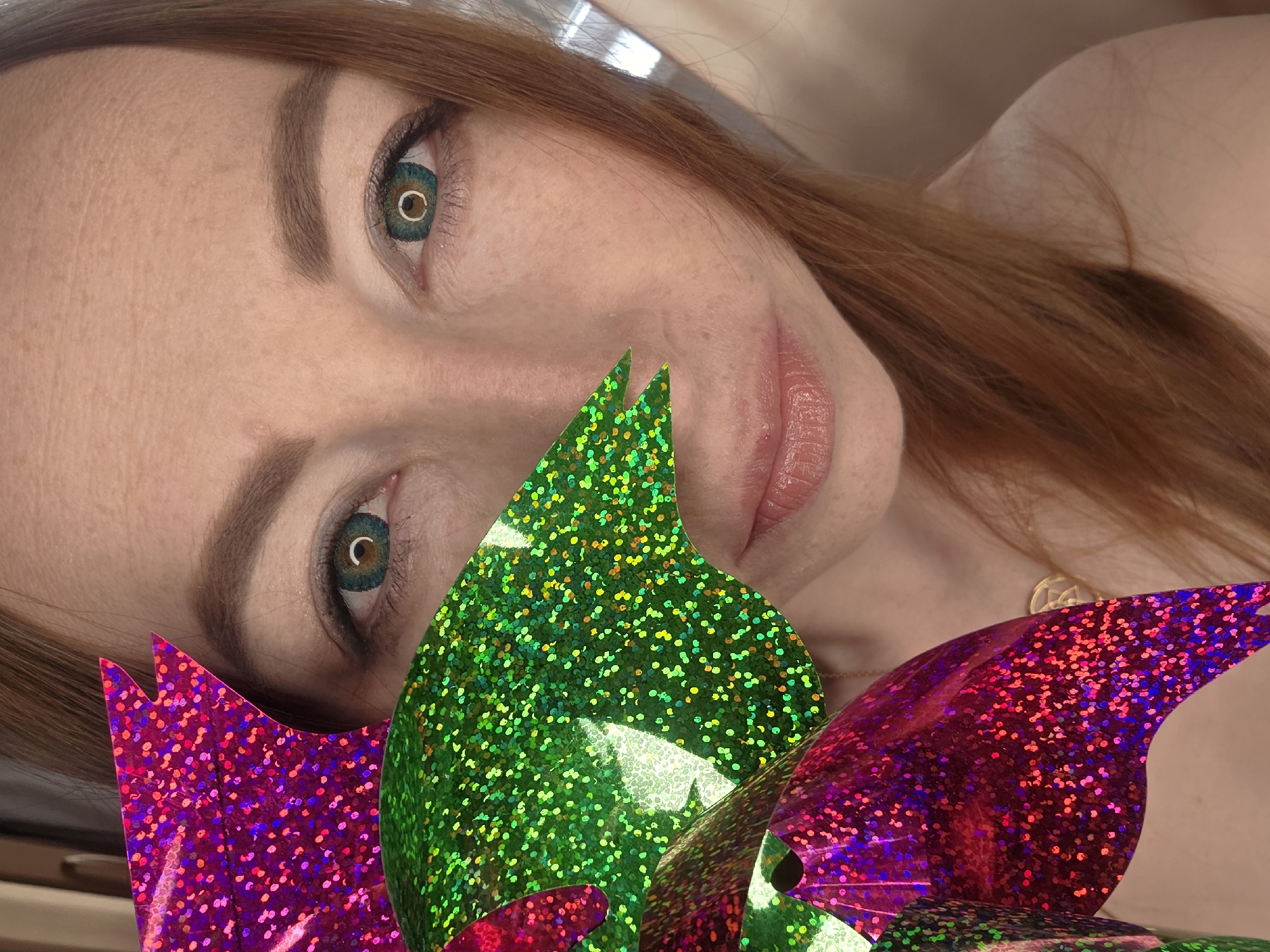 A close-up, angled portrait features a woman with fair skin and long, reddish-brown hair, looking directly at the camera. Her most striking features are her eyes, which are enhanced by prominent green-blue contact lenses with a dark limbal ring and a reflection of a circular light source in the pupils. Her eyebrows are well-defined and dark brown. A subtle, serene expression plays on her lightly glossed, natural-colored lips.
In the foreground, partially obscuring her lower face and shoulder, are two large, shiny, and glittery objects. One is a vibrant green, adorned with green and yellow glitter, and appears on the left side of the frame. The other, positioned to the right, is magenta/purple with red and blue glitter. These objects have a leaf-like or wing-like shape and catch the light, creating a sparkling effect. A delicate gold chain necklace with a small, round pendant is visible on her neck.
The image is a posed photograph, likely taken indoors given the soft, even lighting and lack of outdoor elements. The woman's head is tilted, adding dynamic balance to the composition. The background is out of focus, maintaining emphasis on the subject and the glittery props. There is no visible text in the image. This media file was taken in Chaikovsky, Russia.