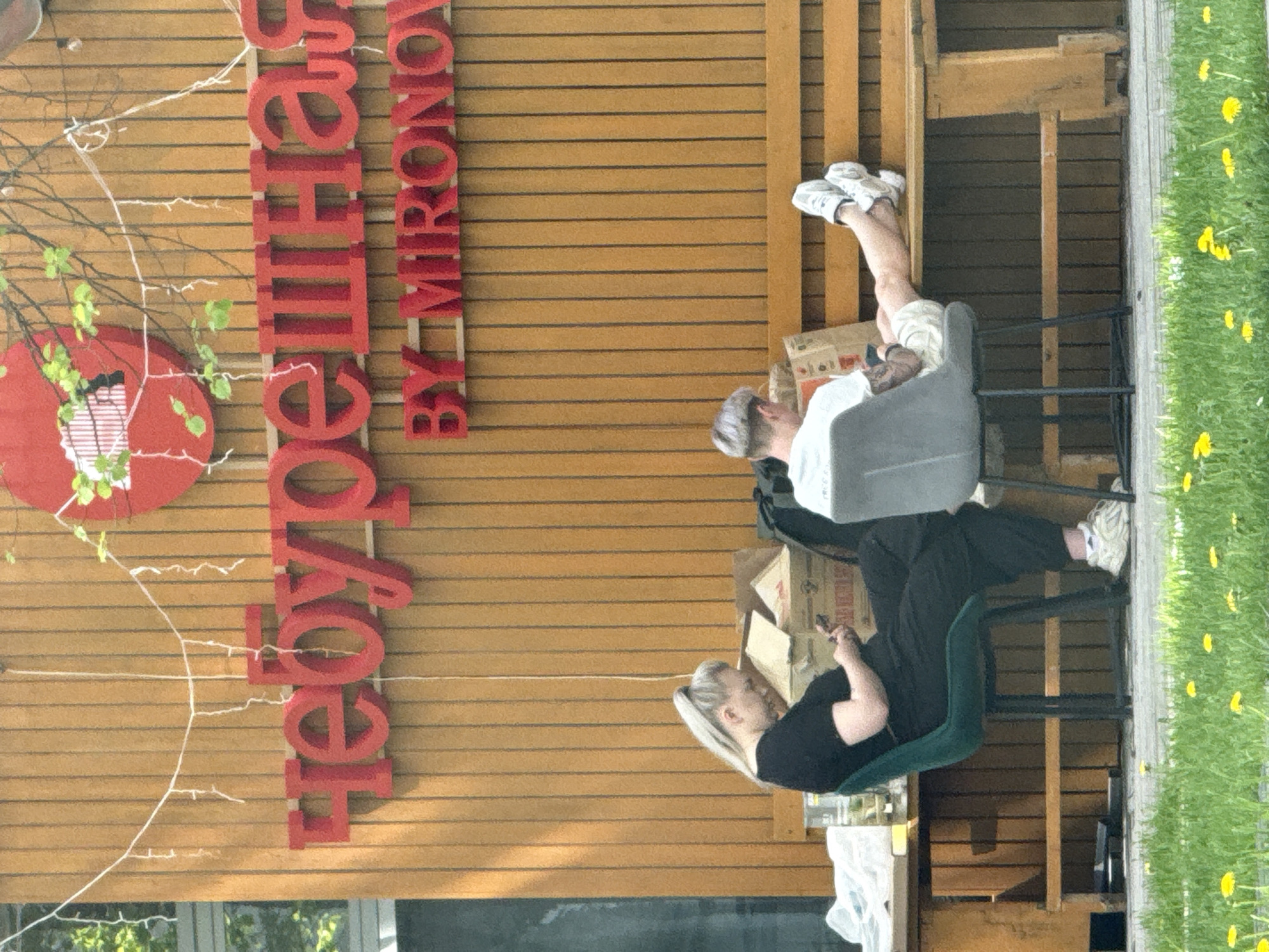 Two people are seated on chairs near a wooden wall bearing the inscription "Чебуречная BY MIRONOV" in red letters. The location is Moscow, Russia. One individual is reclining with their feet elevated, while the other is seated upright, possibly using a mobile device. The setting includes a grassy area with dandelions. Discarded cardboard boxes are also present.