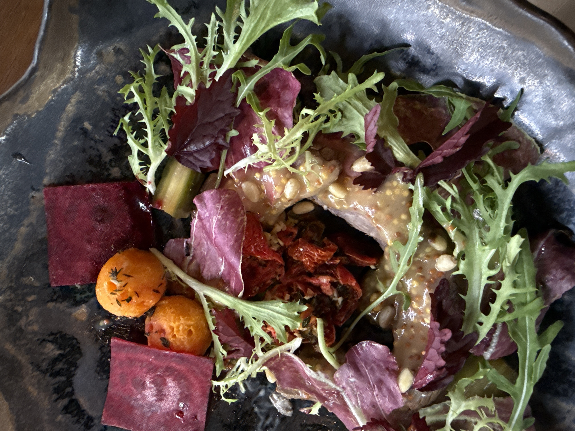 The image shows a salad in a dark, irregularly shaped bowl with a textured, glazed finish. The salad consists of mixed greens, including a variety of leaf lettuce in shades of green and red. There are sliced beets, presented as deep red squares, and two small orange spheres that appear to be sweet potato or carrot balls seasoned with herbs. The salad also contains dried tomatoes and a protein covered in a mustard seed dressing and garnished with pine nuts. The dish is possibly served in a restaurant in Ubud District, Indonesia, as the restaurant identified may have offered a similar dish. The scene is focused on the food itself, highlighting the dish's presentation and ingredients.