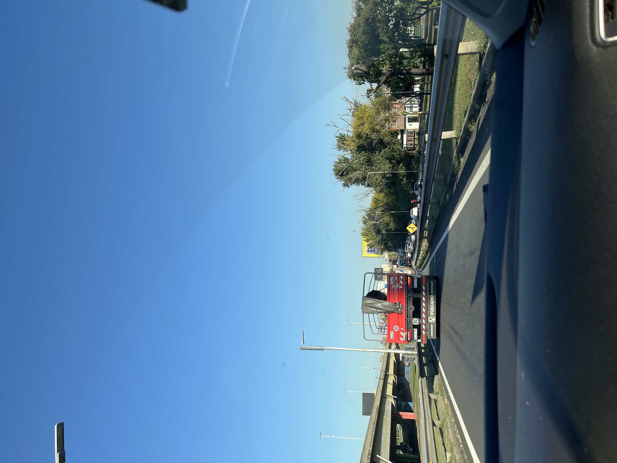 The image, taken from inside a vehicle, shows a view of a highway in Dock Sud, Argentina. A red VULCANO service truck is directly ahead. In the background, there are buildings and trees lining the roadside. A blue sky dominates the upper portion of the image. The image is rotated 90 degrees counterclockwise. The highway appears to be congested with traffic.