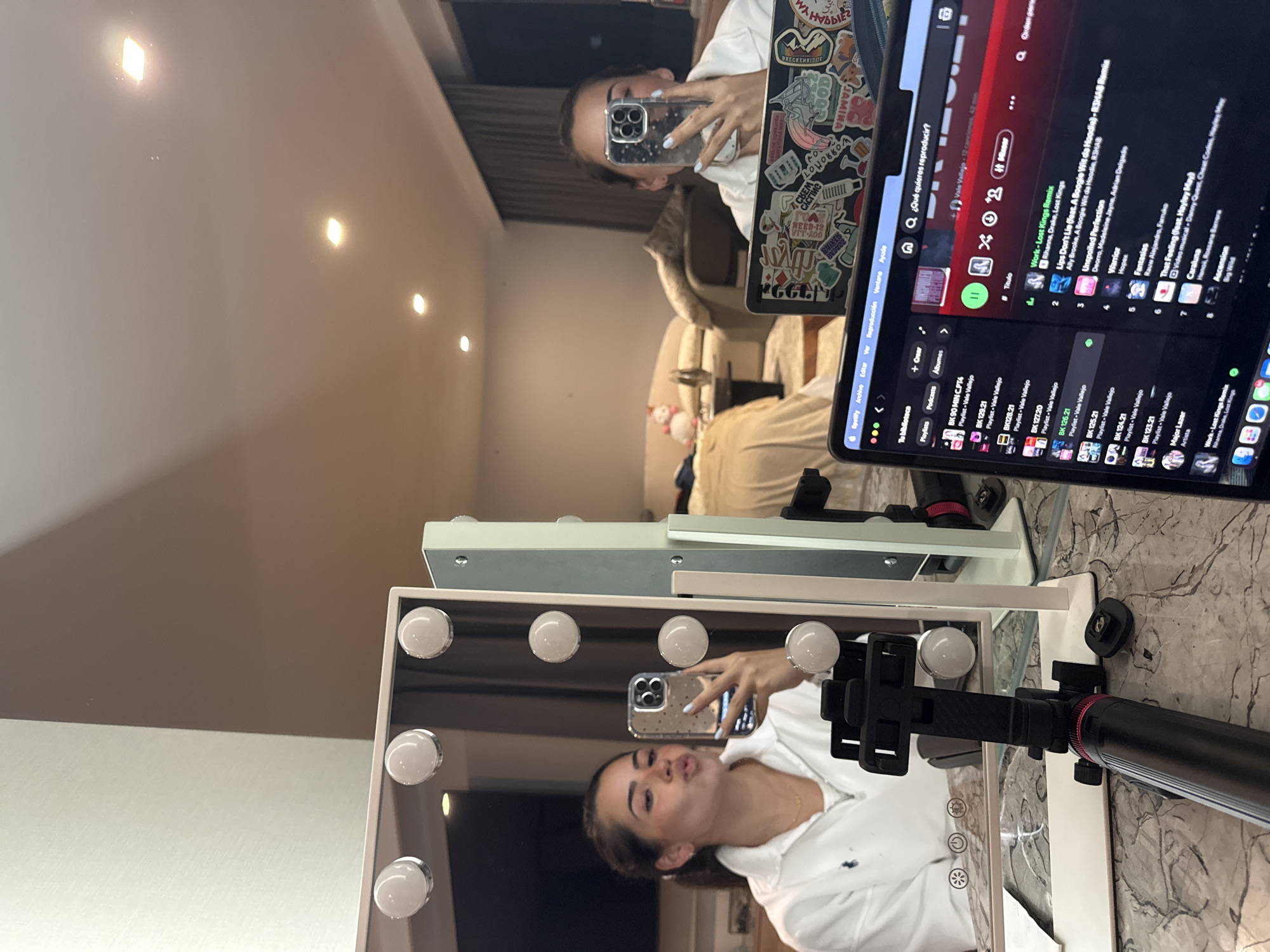 The image shows a reflection of a person taking a selfie with a cell phone in a makeup mirror. The makeup mirror is equipped with surrounding light bulbs. In the background, another person is taking a picture of a laptop with stickers on the back, showing a Spotify interface. The setting appears to be indoors, possibly a bedroom, with a bed and other furniture visible in the reflection. The city and country are not discernible from the visual information available.