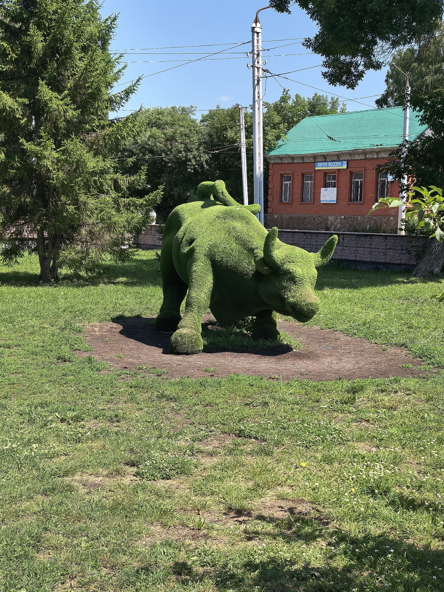 An outdoor, eye-level, medium shot captures a topiary sculpture of a bull in a park on a sunny day. The bull is positioned in the center of the frame, facing right, with its head lowered as if grazing. Its body is meticulously sculpted from green foliage, resembling a large, stylized animal. Behind the bull, a red brick building with a green roof stands under a clear blue sky. The building has several windows and a sign that reads "ПОЧТА РОССИИ," indicating it is a Russian post office. The ground around the bull is covered in a patch of dark soil, surrounded by a grassy lawn with scattered small white flowers. Trees are visible in the background and to the left of the frame, with power lines strung across the upper portion of the image. The lighting suggests it is midday, with distinct shadows cast by the bull and other elements. The overall scene conveys a sense of public space, likely a park or a square, in a Russian city, potentially Sterlitamak given the presence of a Russian post office sign.