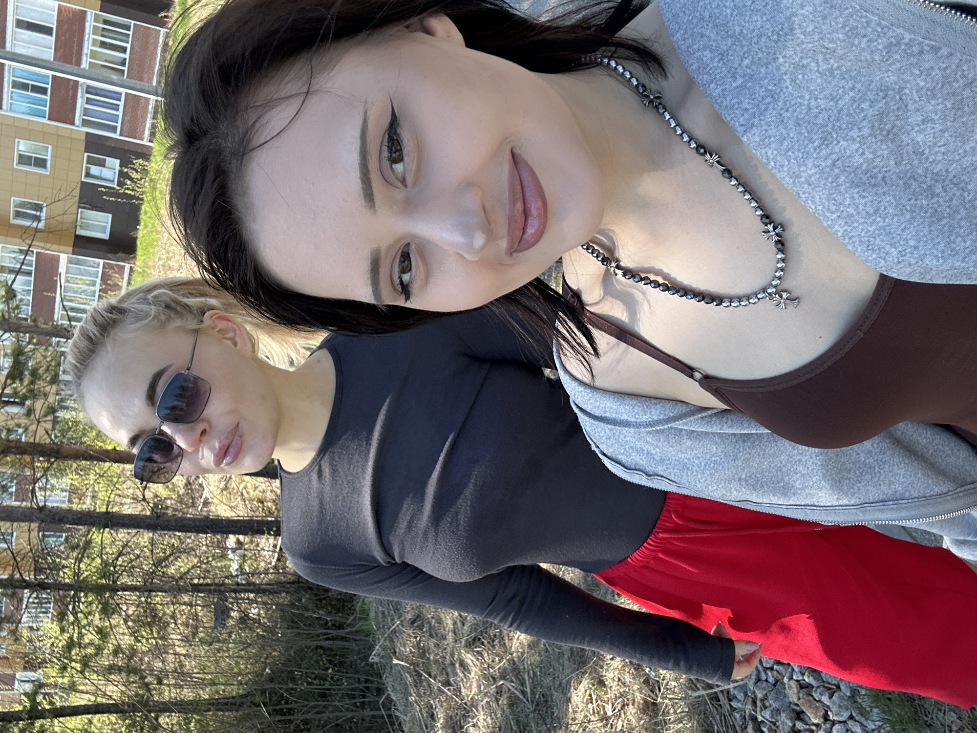 Two young women are posing for a selfie outdoors. One woman has dark hair, brown eyes, and is wearing a brown top, gray zip-up hoodie, and a silver necklace with cross charms. The other woman has blonde hair, is wearing sunglasses, a long-sleeved black top, and red pants. They are standing in a grassy area near trees. In the background, a building with multiple windows can be seen. The location is Высокий, Russia.
