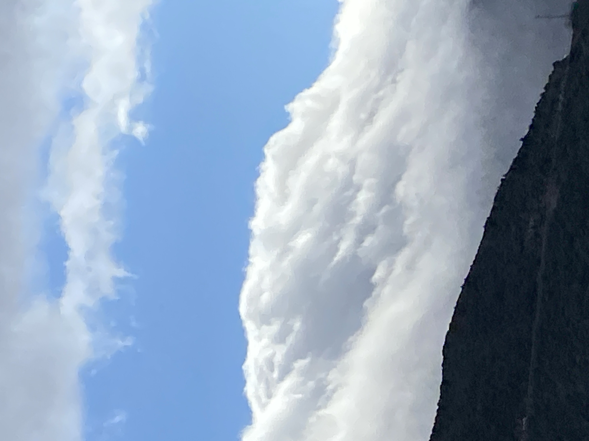 The image displays a daytime landscape scene in Budva, Montenegro, characterized by a mountainous terrain partially enveloped by clouds. The upper third of the frame shows a bright blue sky with two narrow, scattered white cloud formations. Below this, a large, dense formation of white, billowy cumulus clouds dominates the middle section, appearing to crest over a dark mountain ridge. The clouds exhibit distinct, brightly lit edges and varying levels of translucency. The lower third of the image features a dark, rugged mountain slope, appearing to be covered in sparse vegetation, extending from the lower-left corner upwards towards the right. A faint, vertical structure, possibly a telecommunications tower, is visible on the lower-right slope of the mountain, partially obscured by the cloud cover or atmospheric haze. No individuals or specific actions are depicted.