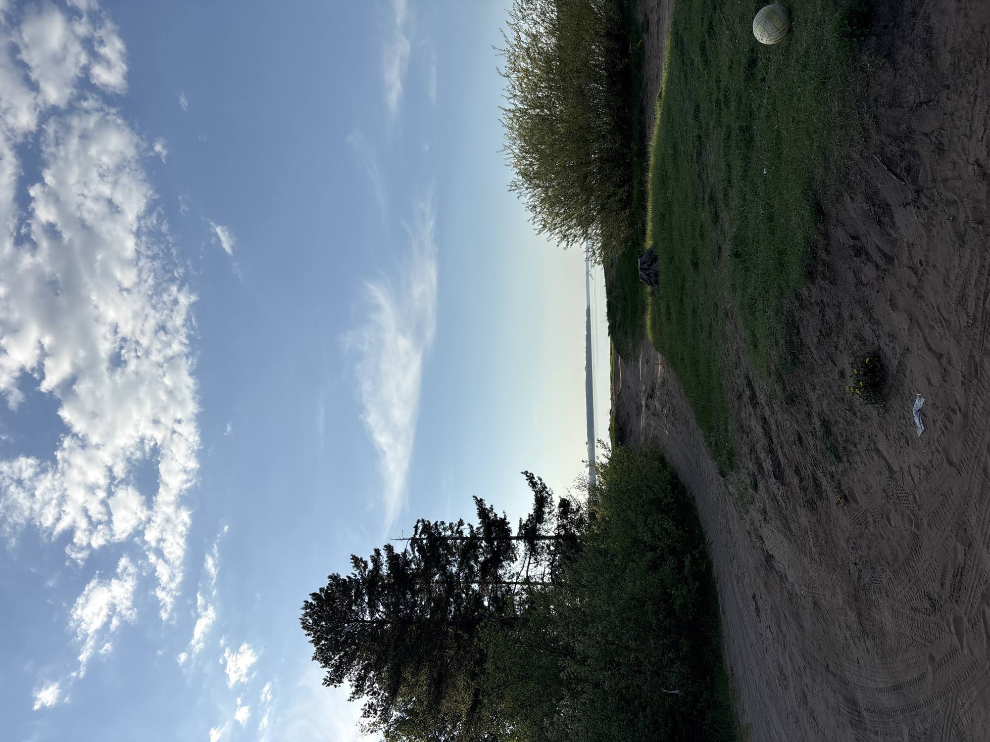 The image shows an outdoor scene in Chaikovsky, Russia. The perspective is oriented sideways, with the sky dominating the left half, featuring a blue sky with scattered white clouds. The right half depicts a dirt path winding through grassy areas and vegetation.
A large, spherical object, possibly a ball, rests on the grass in the upper right corner. Along the path, there are various tire tracks imprinted in the dirt, suggesting vehicular traffic. Scattered pieces of litter are visible.
Different types of trees and shrubs line the path. A tall, coniferous tree is located on the bottom left. Further in the background, there is a view of the Kama River.