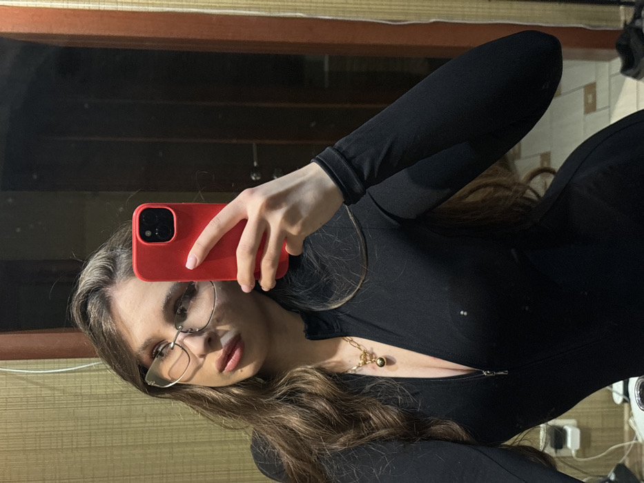 A woman with long, wavy brown hair poses for a mirror selfie. She wears wire-rimmed glasses and a form-fitting black outfit with a slight zip neckline, accessorized with a gold necklace. She holds a red smartphone up to her face. The reflection shows the room behind her including dark wooden trim around the mirror and a glimpse of wall outlets and a white appliance.