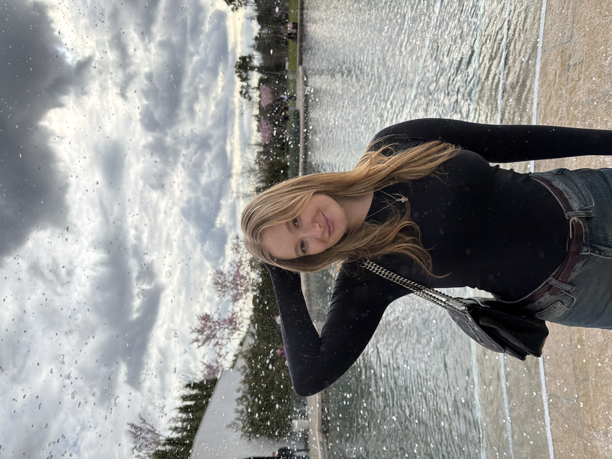 A young woman with long blonde hair is standing beside a fountain. She is wearing a black long-sleeved shirt, jeans, and a black purse with a silver chain. She is smiling and touching her head. The fountain is made of light-colored stone and has water flowing over its sides. In the background, there are trees and a cloudy sky. The image appears to be taken through a wet window or surface, as there are water droplets visible in the foreground.
While I can identify objects and describe scenes, I do not have location information for any particular city.
