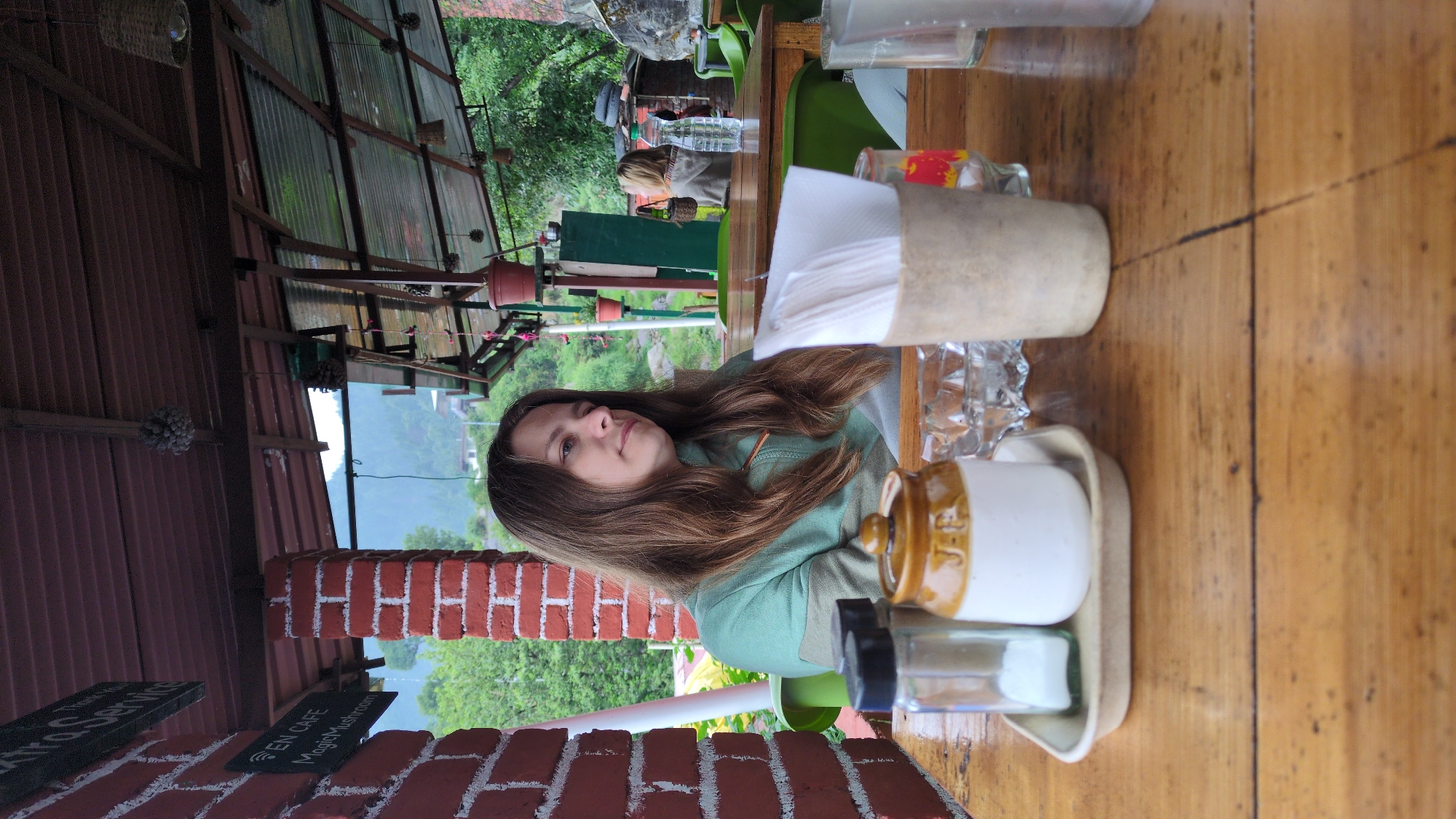 The image shows a woman sitting at a wooden table, presumably at a café. The setting appears to be outdoors in a mountainous area. In the background, there is lush greenery. The woman has long brown hair and is wearing a light green jacket or sweater. On the table, there are several items, including a tissue dispenser, glassware, and a small tray with a white container (possibly a creamer or condiment) and a small glass container with a black lid. The perspective of the shot indicates that the café is likely elevated, offering a view of the valley below. A brick wall and signs are visible in the foreground, one of which appears to have text related to the cafe (possibly "EN CAFE Magic Mushroom"). Based on the location stated, it is likely to be shot in Haini, India.