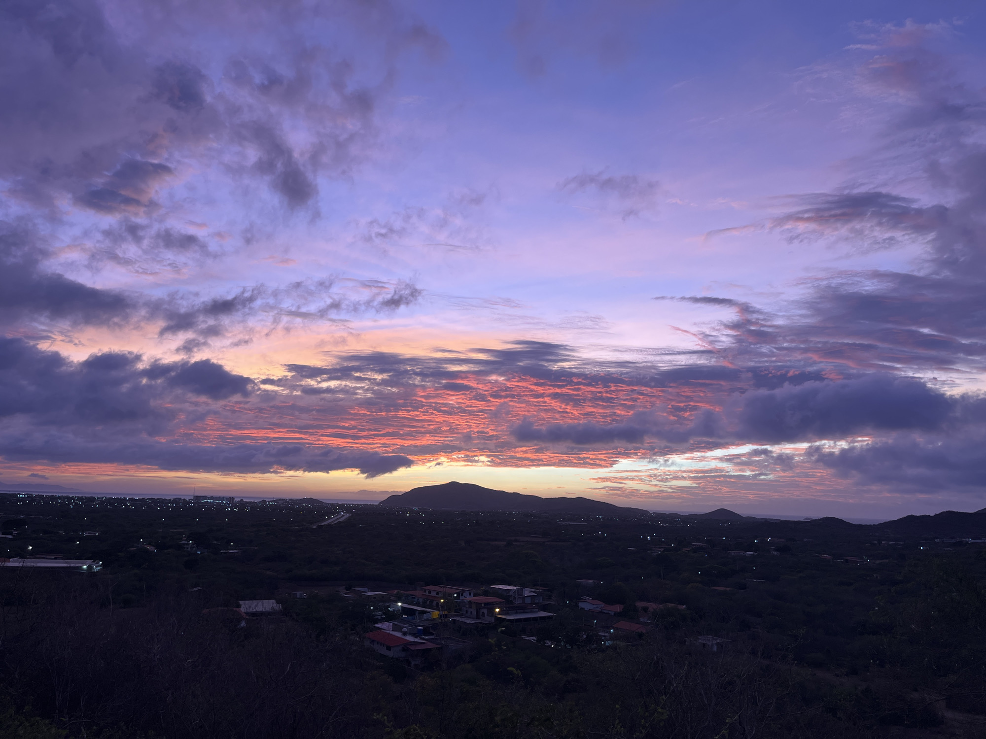 This image captures a twilight scene over a hilly, semi-arid landscape near Santa Ana, Venezuela. The sky is a dramatic canvas of purple, pink, and orange hues, with streaks of vibrant coral coloring the underside of the clouds. Dark, silhouetted hills form the horizon, behind which the faint lights of a town or city are visible. The foreground is dominated by dark, dense vegetation, with a few scattered buildings with red roofs nestled within the landscape. The overall impression is one of a serene, natural setting transitioning from day to night, with human habitation subtly integrated into the environment.