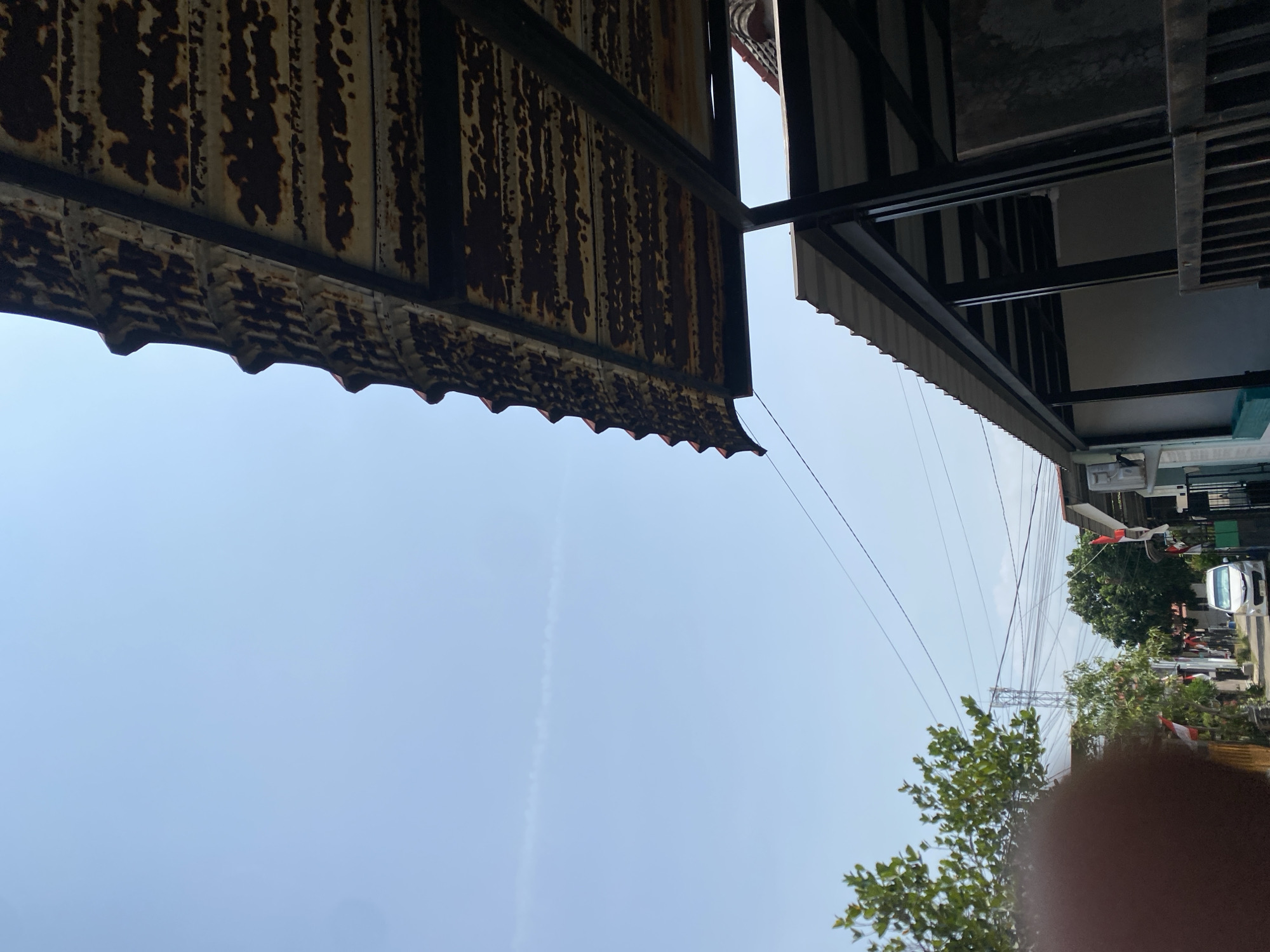 A low-angle outdoor shot captures a clear blue sky with a faint contrail. In the foreground, the weathered, rusty corrugated metal roof of a structure extends into the frame from the right. Below this, another structure with a dark metal frame and a corrugated metal roof is visible. To the left, a narrow street in what appears to be a residential area is lined with trees and houses. Several Indonesian flags are displayed along the street. A white car is parked on the street, and a tall communication tower can be seen in the distance. The overall environment suggests a sunny day in a developed area.