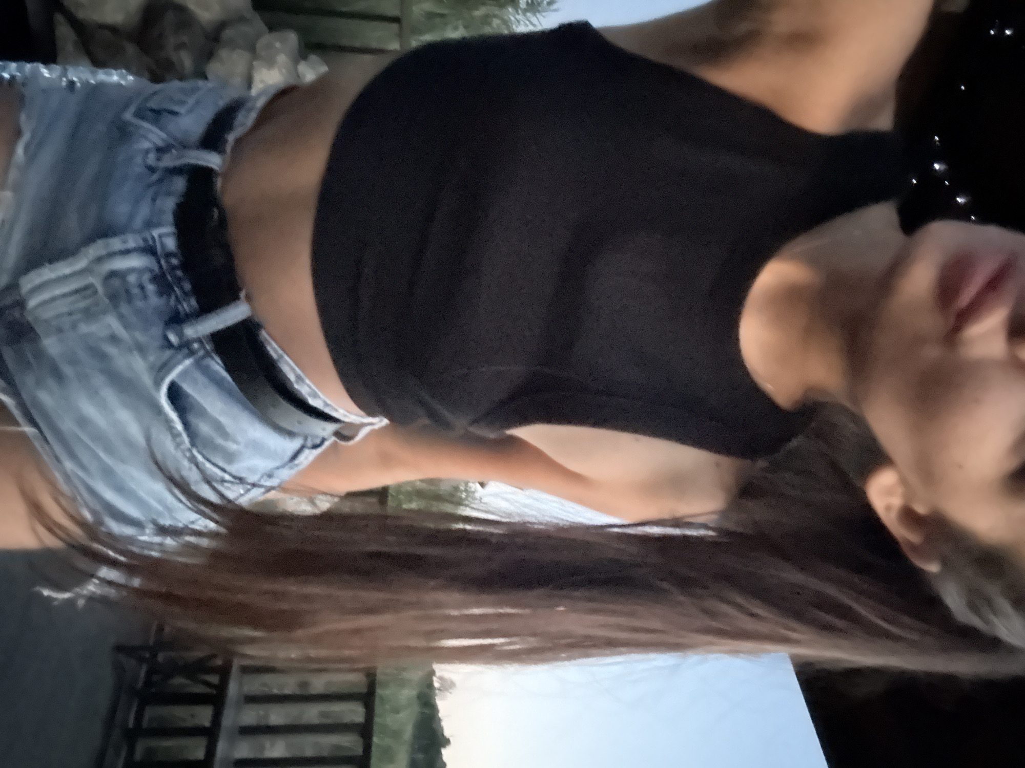 This media file depicts a person, likely female, outdoors. She is wearing denim shorts and a dark, form-fitting top. Her long, dark hair is flowing down, obscuring much of her body. The background includes some rocks and foliage, suggesting a natural or semi-natural setting. There are also hints of man-made structures, possibly a fence or railing, and a glimpse of what appears to be a light-colored sky, potentially indicating dusk or dawn. The overall lighting suggests it is either early morning or late evening. No text is visible.