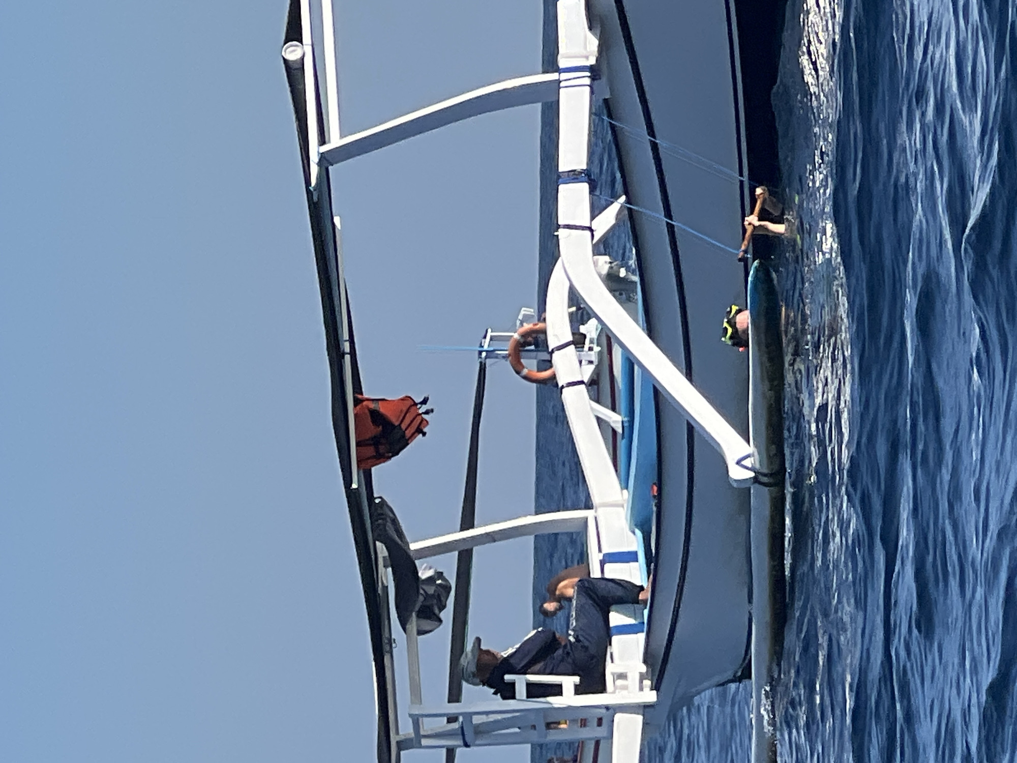 An image captures a clear blue sky above a serene blue ocean, with a traditional Indonesian fishing boat as the central subject. The boat, white with dark trim, features outriggers and a canopy supported by white poles. An orange life vest is suspended from the canopy. On the boat's deck, a person wearing a hat and dark clothing sits with their arms crossed. In the water beside the boat, two individuals are partially submerged, one wearing a snorkeling mask and holding onto the boat. The scene appears to be at sea, with no specific city landmarks identifiable.