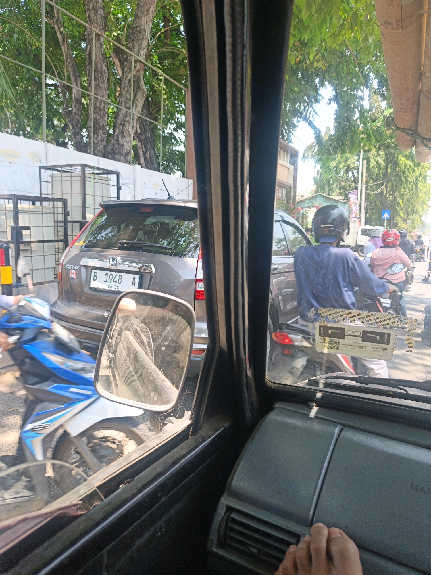 The image is taken from inside a vehicle, looking out at a busy street scene in North Jakarta, Indonesia. The perspective suggests the vehicle is stopped in traffic. In the foreground, part of the interior of the vehicle is visible, including the dashboard and a side mirror. The side mirror reflects a portion of the street, showing what appears to be a motorbike.
Outside the vehicle, a brown Honda CRV is prominently visible, with its license plate reading "B 1948 A" and "06-30". To the left of the CRV, a blue and white motorbike is stopped. Further down the street, several other motorbikes and a few cars are visible, indicating traffic congestion. People are seen riding the motorbikes, most wearing helmets. One person in the foreground, on a motorbike to the right of the CRV, is wearing a dark blue shirt, and another in front of them is wearing a pink jacket and helmet.
The environment appears to be urban, with trees lining the street and buildings in the background. The sunlight suggests it is daytime. The presence of traffic signals and the overall scene are consistent with a typical street in a city like North Jakarta.