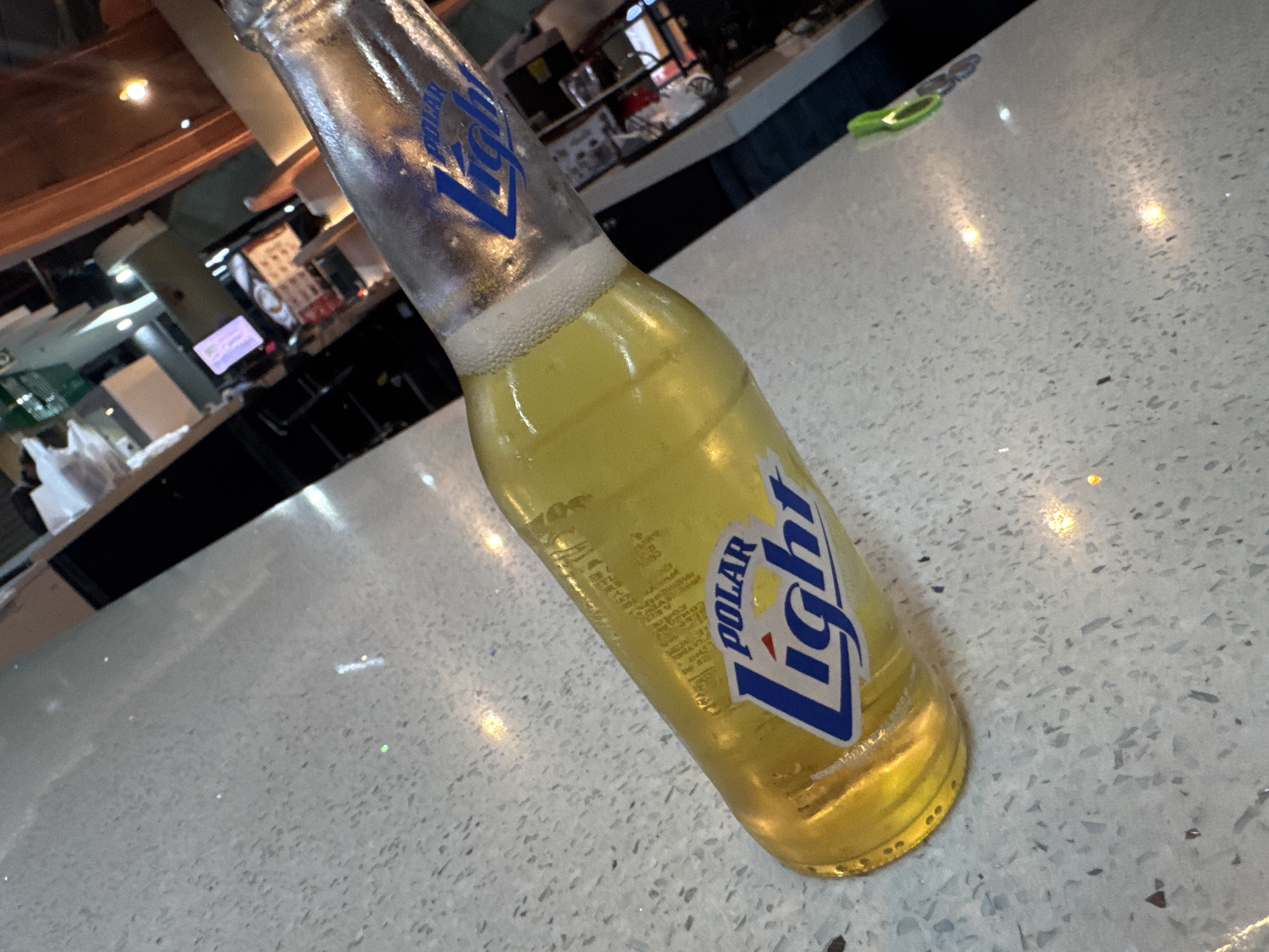 The image shows a partially consumed bottle of Polar Light beer resting on a light-colored, textured countertop. The bottle is positioned diagonally across the frame, with the label clearly visible. The beer is a pale yellow color with a foamy head. In the blurry background, one can discern elements of what appears to be a restaurant or bar setting, including counters, lighting fixtures, and possibly other patrons. The lighting is somewhat dim, creating reflections on the countertop. There is no specific information in the image to definitively determine the location is in Caracas, Venezuela. The image does not depict any people.