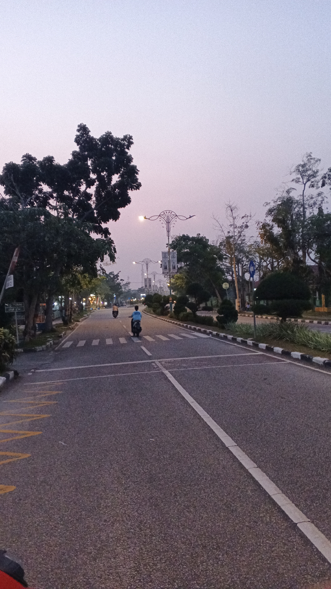 The image depicts a wide asphalt road in Siak Sri Indrapura, Indonesia, possibly near the Siak Riverfront, given the presence of palm trees and lush vegetation lining the street. The sky is a pale, hazy purple, indicative of early morning or late evening. Two individuals are seen riding motorcycles on the road. The person in the foreground, closer to the camera, is wearing a blue shirt and riding a dark-colored motorcycle. Further down the road, another person on a motorcycle is visible. The road is marked with white lines, including a pedestrian crossing with alternating white and black stripes on the edges. Streetlights with ornate designs are intermittently placed along the sides of the road, suggesting a public area. Trees with dense foliage are on the left side of the road, with some smaller, manicured bushes and trees on the right. There are also signs and lampposts visible on both sides of the street. The overall impression is of a quiet, urban street scene during twilight hours.