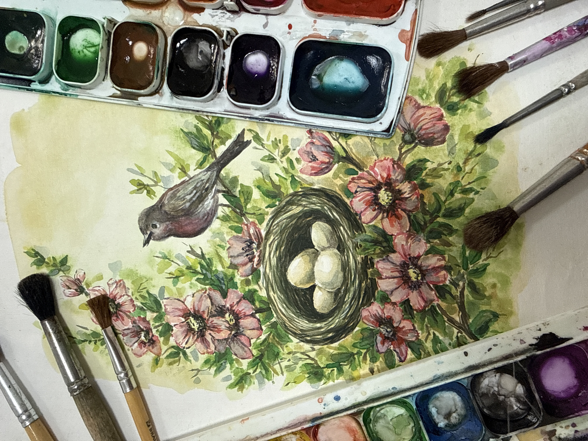 This is a top-down, close-up shot of a watercolor painting in progress, featuring a bird perched on a branch with a nest containing three eggs, surrounded by pink flowers and green foliage. The painting is set against a light yellow watercolor wash. To the top and bottom right of the painting are two white plastic watercolor palettes filled with various colors. Several paintbrushes, with wooden handles and brown bristles, are scattered around the painting and palettes, some resting on the painted surface. The brushes vary in size, with some appearing to be used for finer details and others for broader strokes. The setting appears to be an artist's workspace, likely indoors given the lighting. The time of day and weather are not discernible from the image. No visible text is present on the painting or the artistic supplies. The overall impression is one of creative activity.