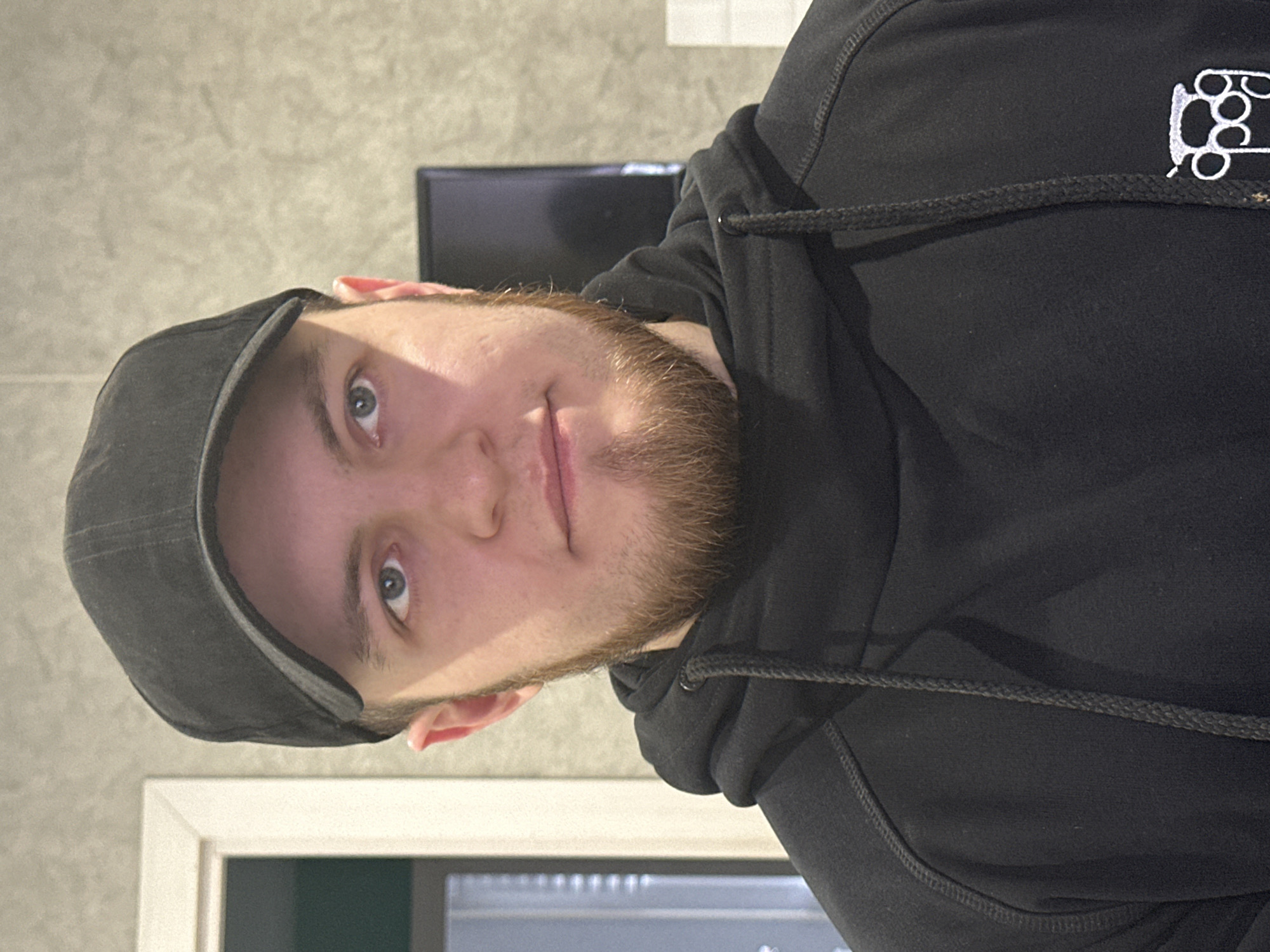 A man is wearing a black newsboy cap and a black hoodie. He has a beard and light blue eyes. The background includes a gray wall, a black screen, and a white doorframe. The man appears to be indoors.
