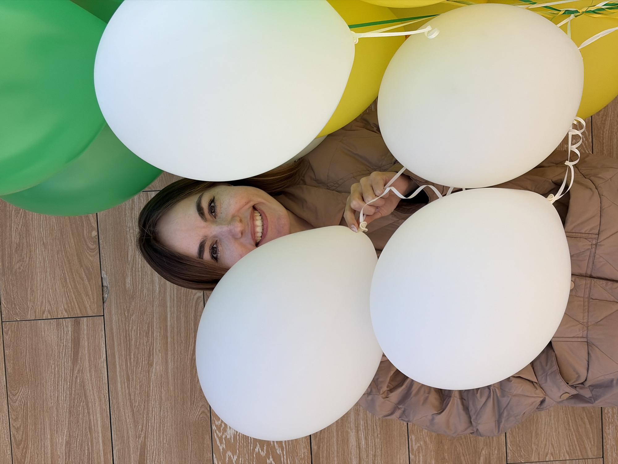 A medium, high-angle shot depicts a smiling woman surrounded by balloons. The balloons are in shades of white, green, and yellow. The woman has light brown hair and is wearing a tan-colored jacket. The background is a wood-patterned floor. The location is potentially Izhevsk, Russia.
