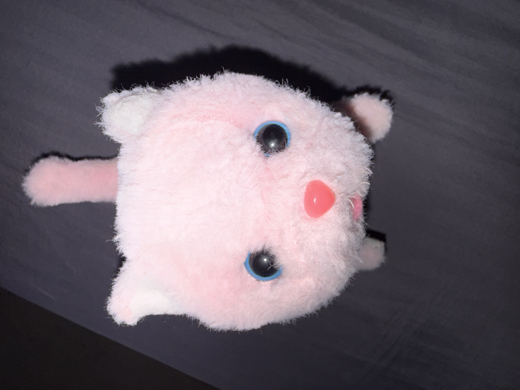 This is a close-up, overhead shot of a pink, plush cat toy with large blue eyes and a pink heart-shaped nose. The toy is positioned on a dark, textured surface that appears to be fabric. The lighting is somewhat dim, casting a shadow to the right of the toy. The toy has two pointed ears and a tail extending upwards from its head. There is no indication of the city of Cahul, Moldova, or any contextual information that would place the image within a specific geographical location.