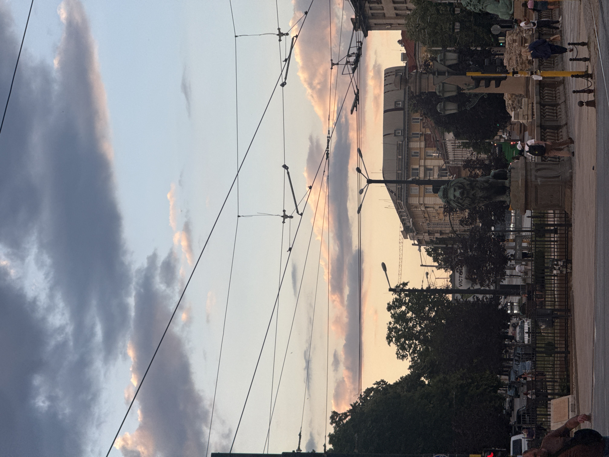 The image captures a street scene in Sofia, Bulgaria, likely during either sunrise or sunset, given the soft, warm hues in the sky and the pale, diffused light. The sky is a canvas of soft blues and pinks, with scattered grey clouds. Overhead, a complex network of tram wires crisscrosses the frame, indicating a city with public transportation.
In the mid-ground, buildings with distinct architectural styles line the street, suggesting an urban environment with a mix of older and possibly more modern structures. A prominent statue of a lion is visible, a significant landmark, possibly the Lion's Bridge or a related monument, given the context of Sofia. People are present, some walking along the sidewalk or crossing the street, and a few are gathered near the statue. Their activities appear casual, typical of a public space. A green bus and other vehicles are also present, suggesting ongoing traffic.
In the background, trees provide a touch of greenery amidst the urban landscape. A street lamp is also visible. There is no discernible text that is legible in the image. The overall mood is peaceful and perhaps a bit tranquil, characteristic of the transitional light of dawn or dusk in a European city. The orientation of the image is rotated 90 degrees clockwise.