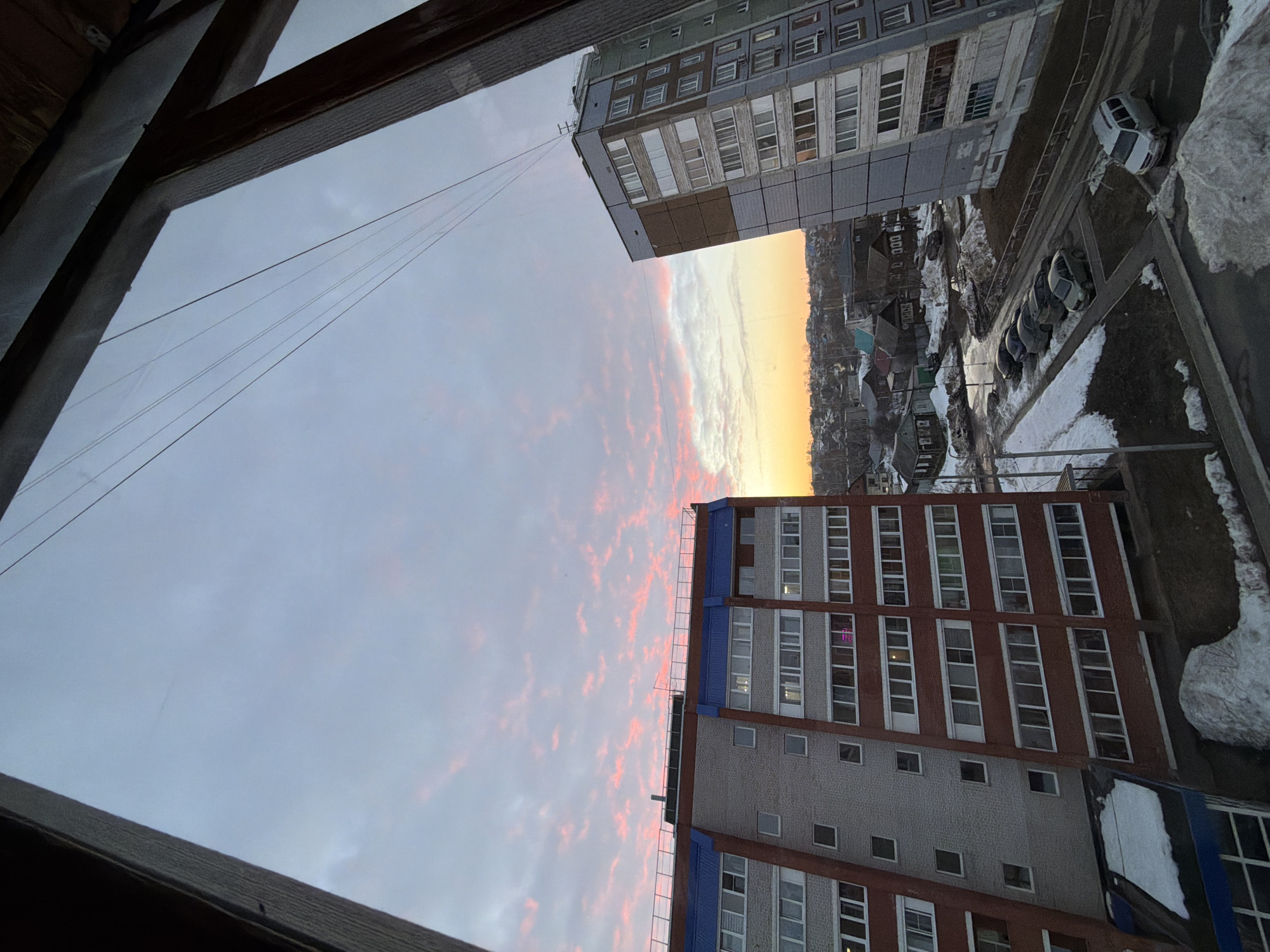 This image presents an elevated view within the city of Izhevsk, Russia, captured during twilight hours. The sky exhibits a gradient, transitioning from a light blue-grey overhead to vivid pink and orange cloud formations along the horizon, indicative of either sunrise or sunset. Several thin, dark utility lines are visible against the sky in the upper right quadrant. The scene is framed on the left, right, and top by dark, rough-textured window sills and frames.
Two prominent multi-story residential buildings anchor the scene. On the left, a structure approximately 9-10 stories tall features a lower facade of reddish-brown brick and an upper section of light grey brick, topped with a distinct blue roof section and a fenced flat roof. On the right, another similar-height residential building displays a facade composed of light grey and brown panels. Both buildings contain numerous rectangular windows, some illuminating interior spaces.
The ground level between these structures and in the immediate foreground consists of paved areas, including a parking lot. These surfaces are marked by significant patches of dirty, melting snow, areas of slush, and standing water, indicating thawing conditions. Several vehicles are present: a silver minivan is parked diagonally in the lower right, and a line of approximately five to six passenger cars (including grey, dark blue, and black models) are parked perpendicular to a curb further back.
Beyond the foreground buildings, the urban landscape extends with a dense concentration of lower-rise, older residential houses, many constructed of wood, featuring varied roof colors such as blue and green. Bare deciduous trees are interspersed throughout this background. No human subjects are visible within the frame.